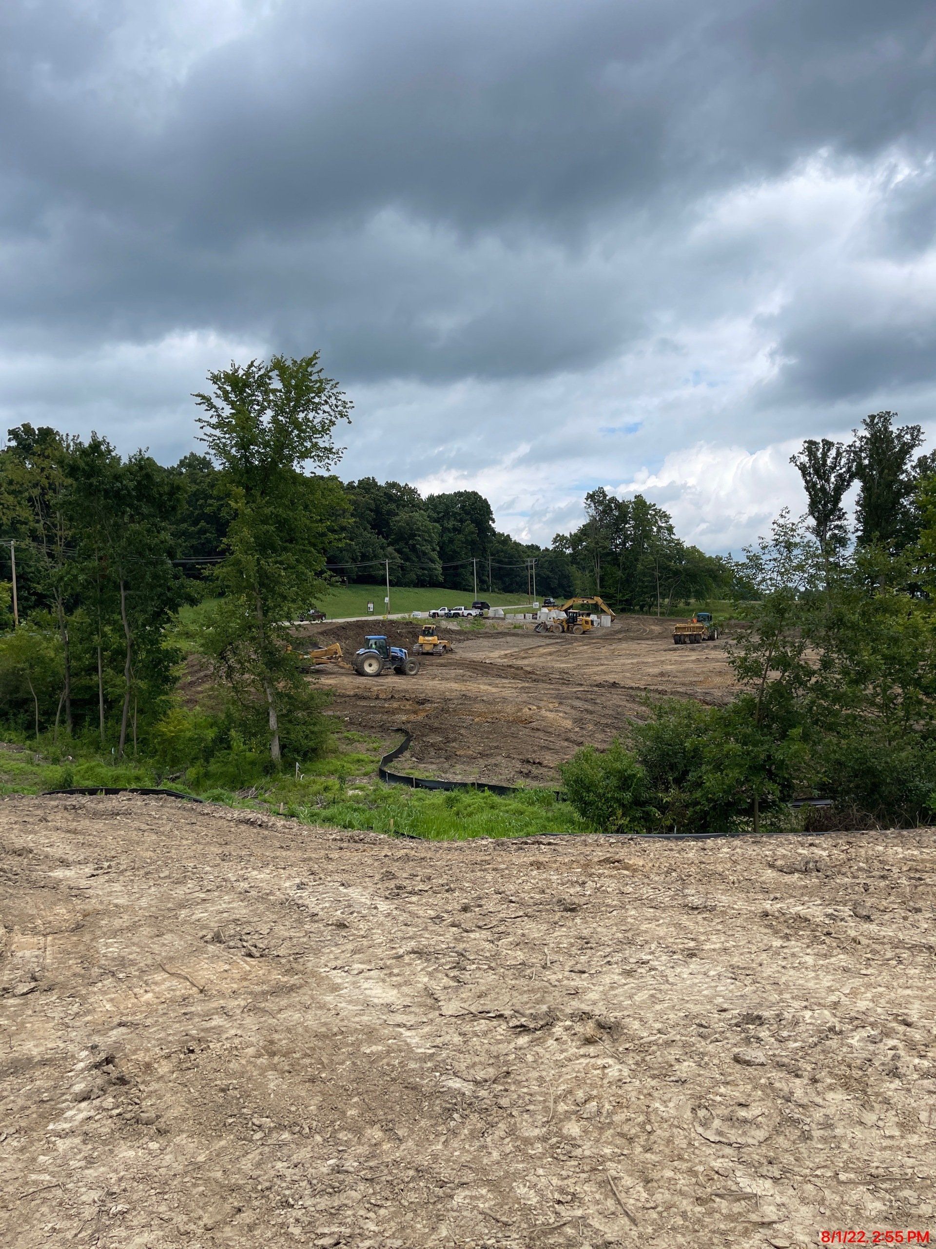 Construction site with earthmovers clearing land under a cloudy sky. Trees frame the edges.