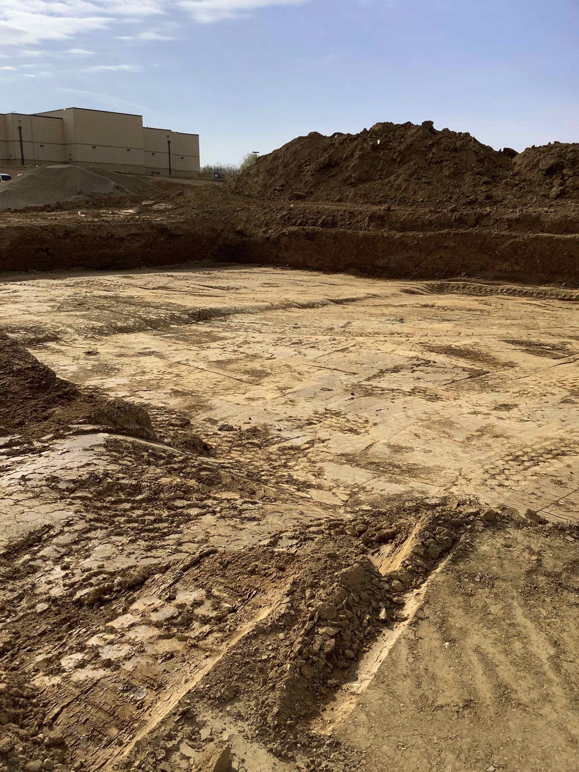 Construction site: Dirt ground with tire tracks, large pile of dirt, and a building in the background.