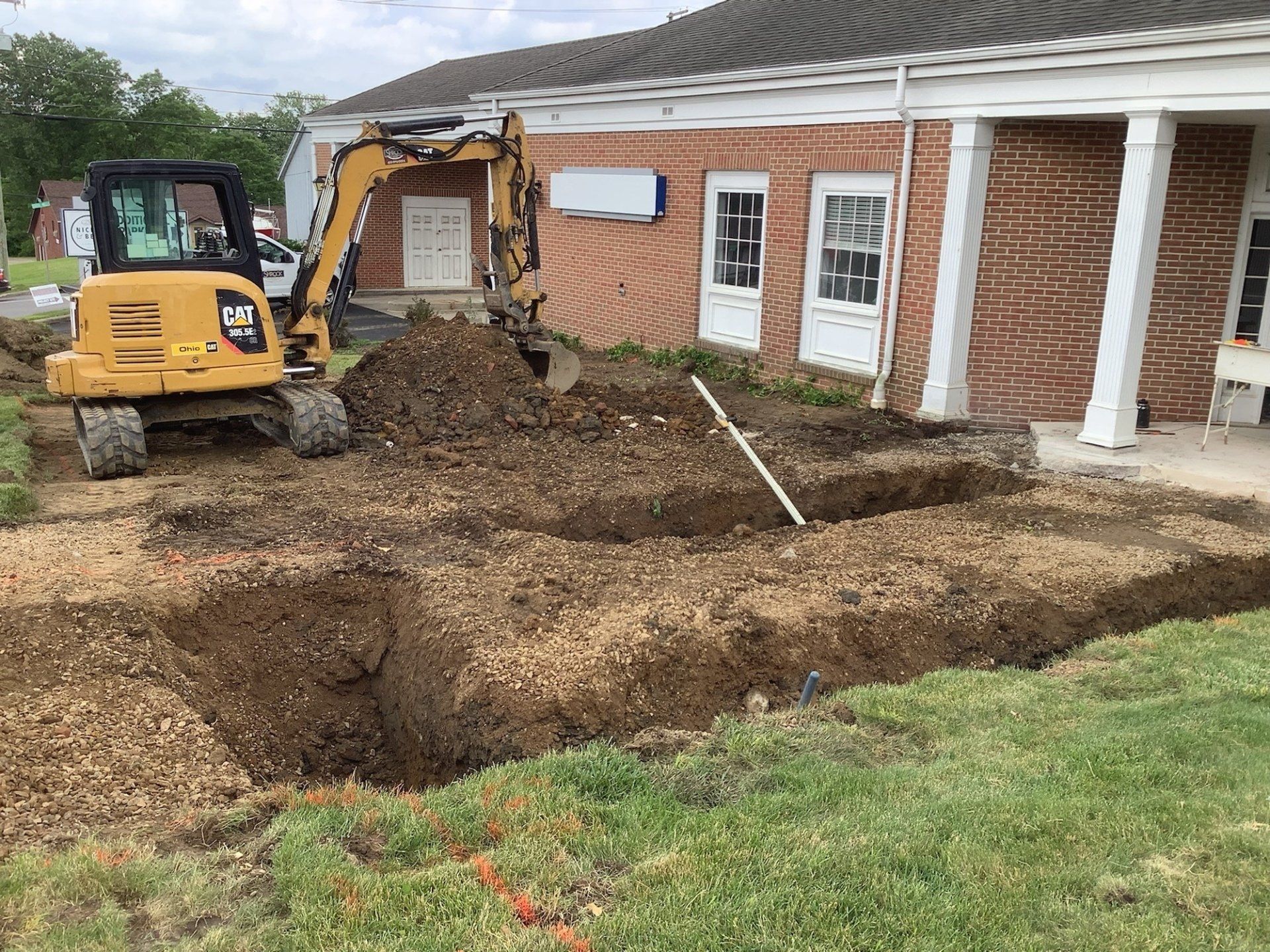 Yellow excavator digging near a brick building with white columns; soil and gravel in the trench.