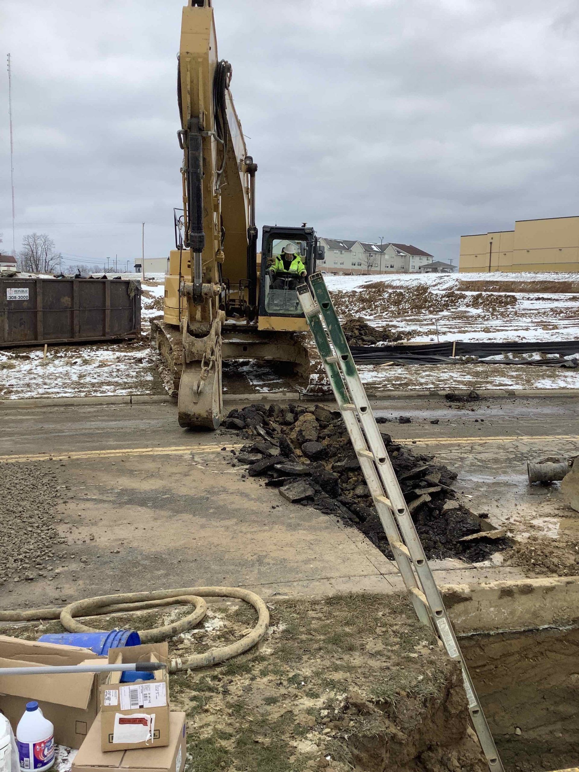 Excavator digging a trench on a construction site; a ladder extends into the trench.
