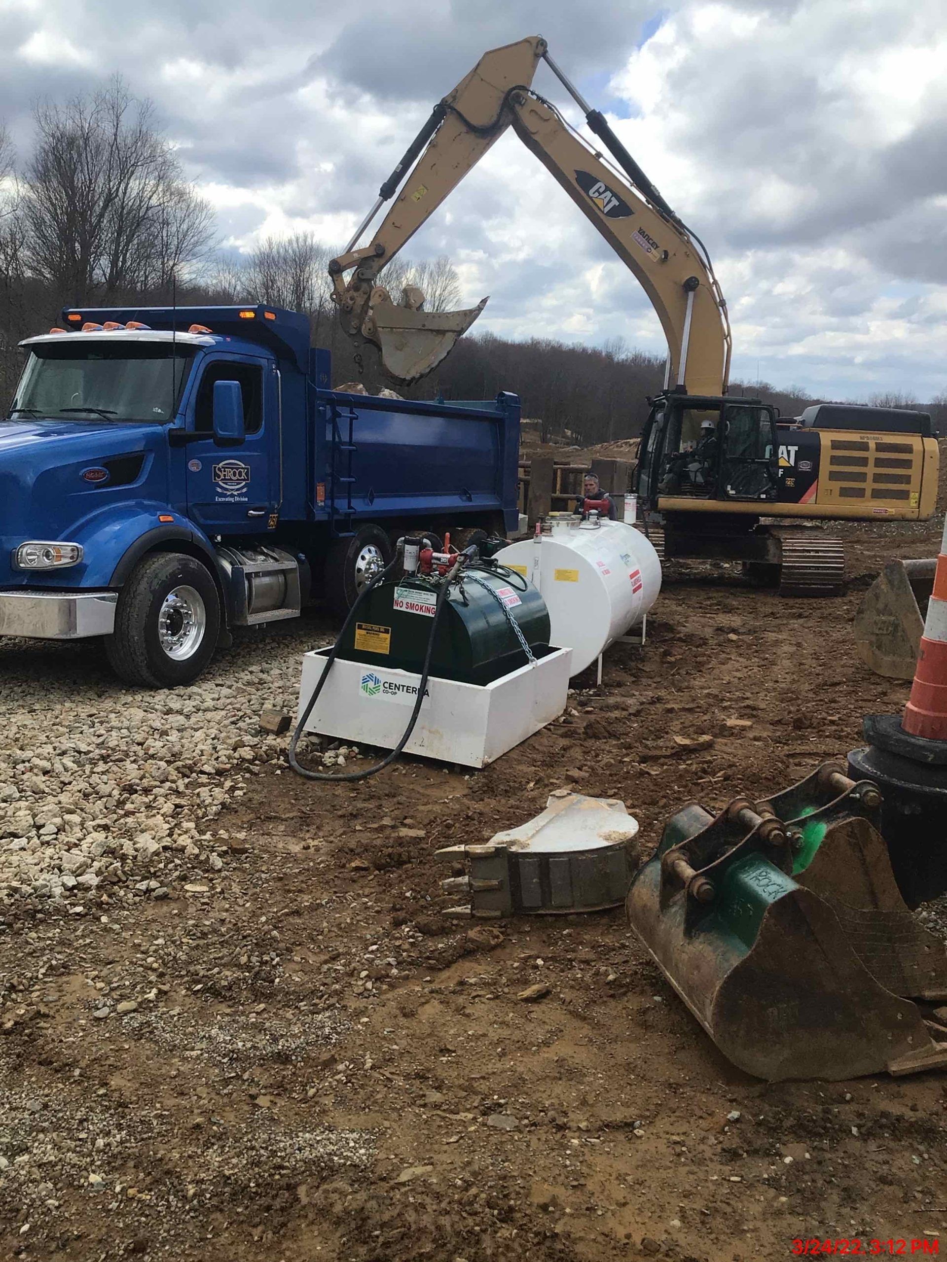 Construction site: excavator loading truck, fuel tank, and equipment. Blue truck, yellow excavator, dirt.
