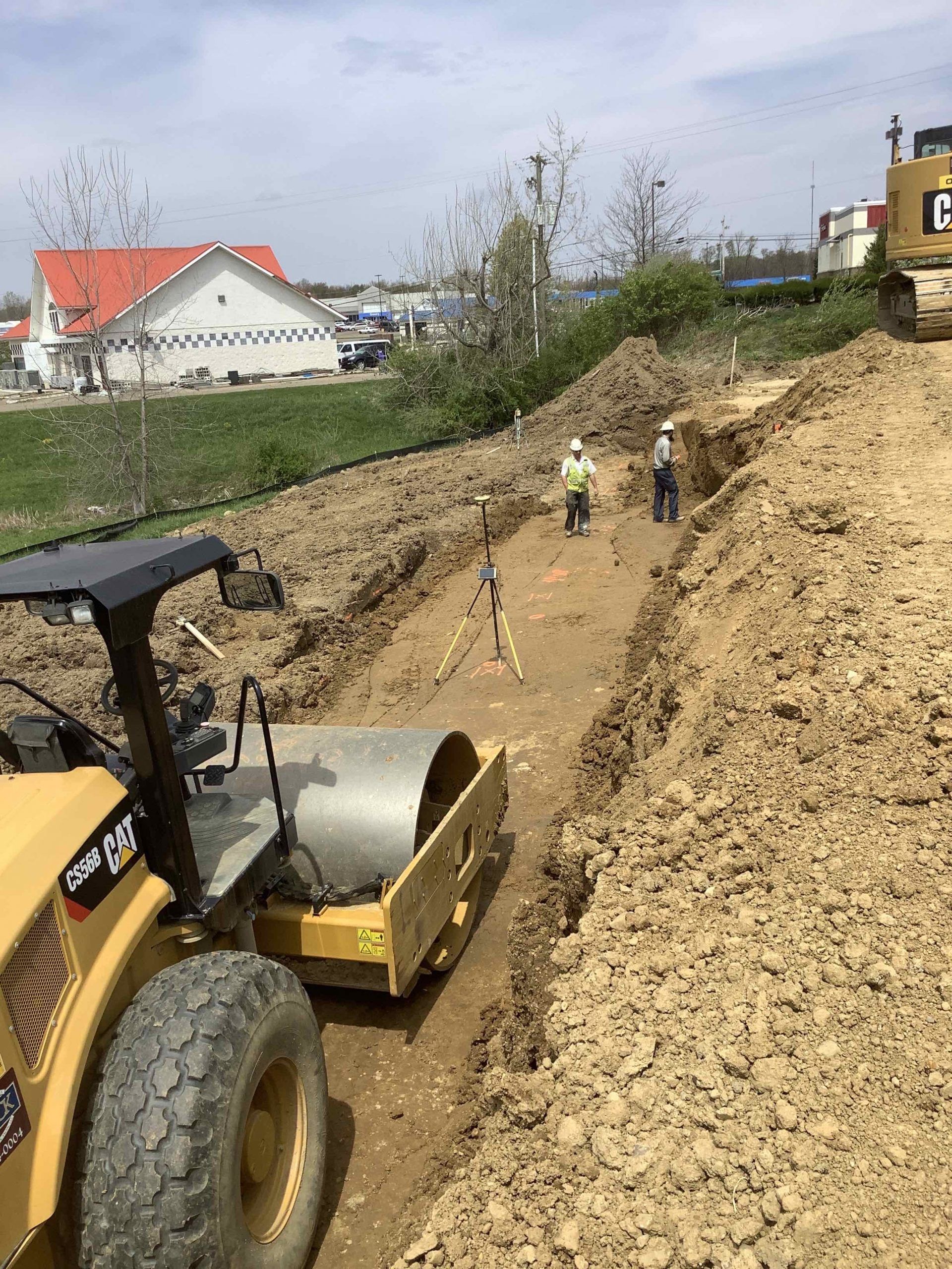 Construction site: Roller compacting trench. Two workers, equipment, and buildings in background.