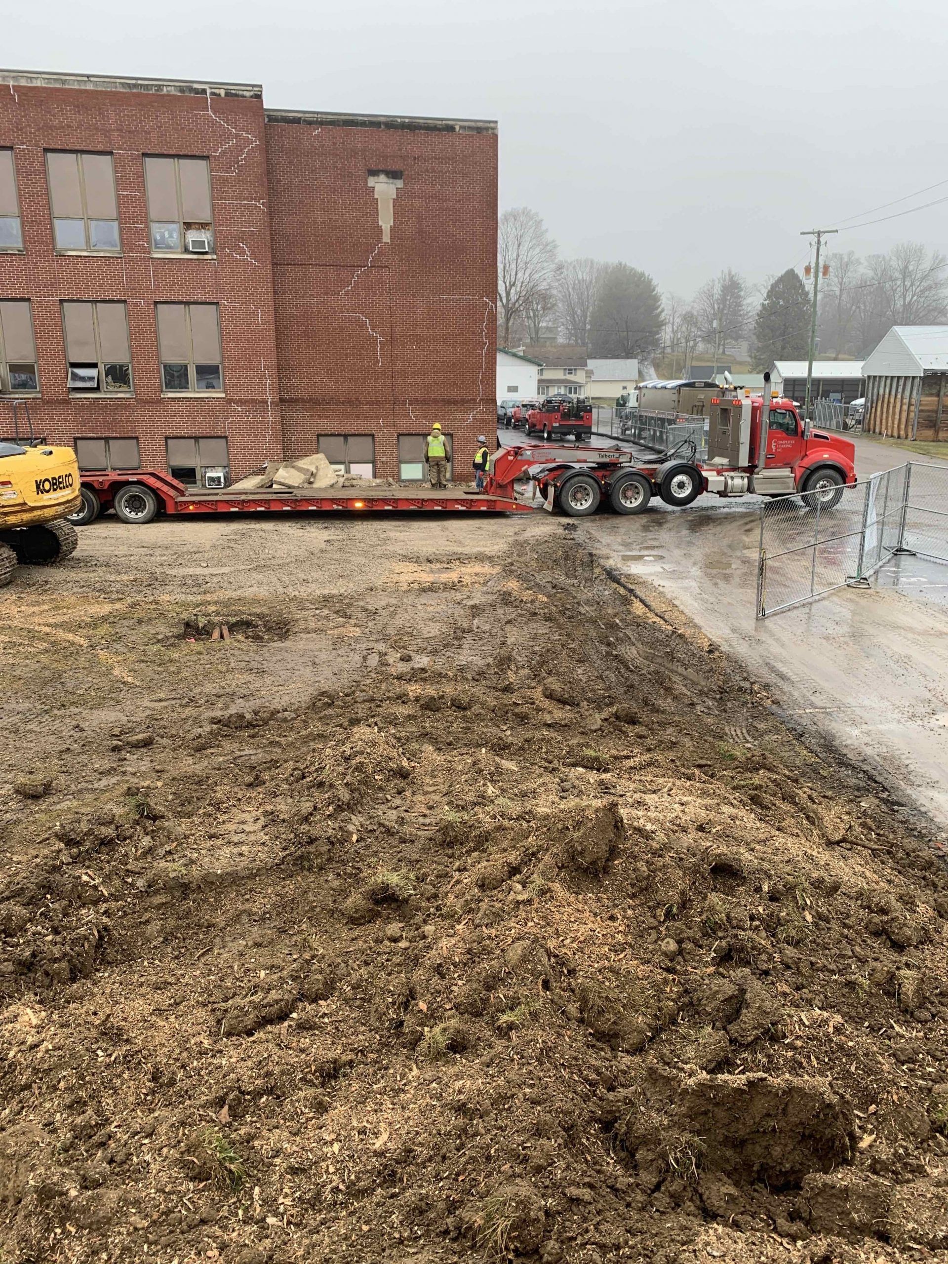 Truck hauling equipment near a brick building on a muddy construction site.