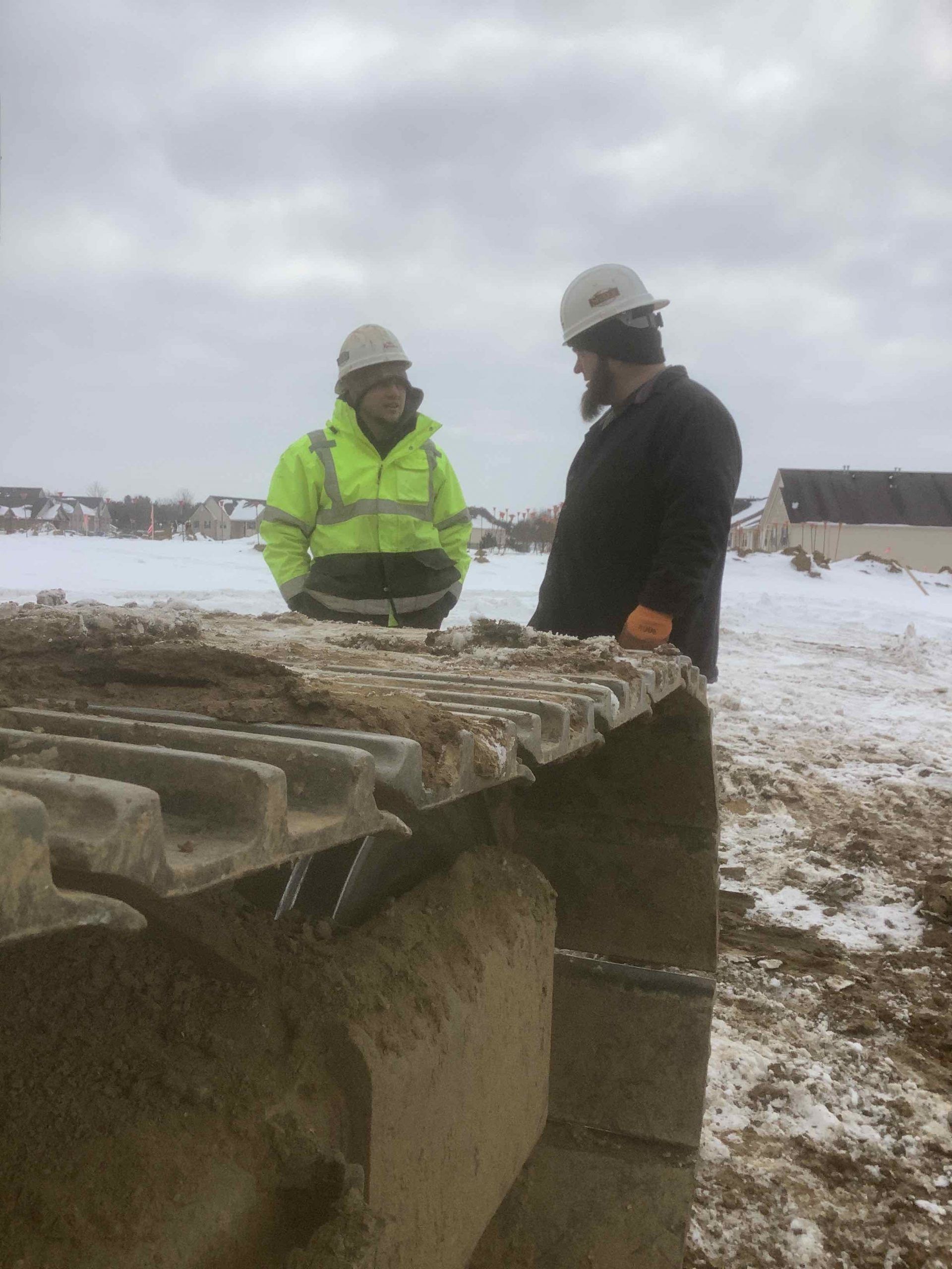 Two construction workers in hard hats converse near a tractor track on a snowy construction site.