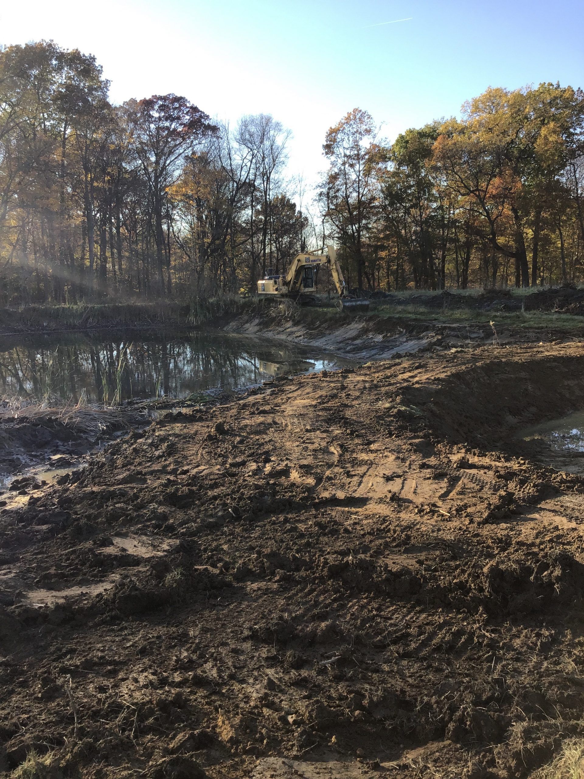 Muddy ground with standing water, a construction site with trees in the background under a sunny sky.