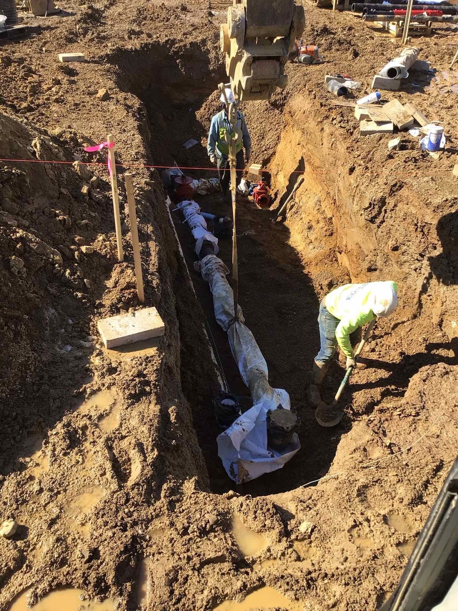 Construction workers in a trench; an excavator lifts a large pipe. Brown dirt surrounds the trench.