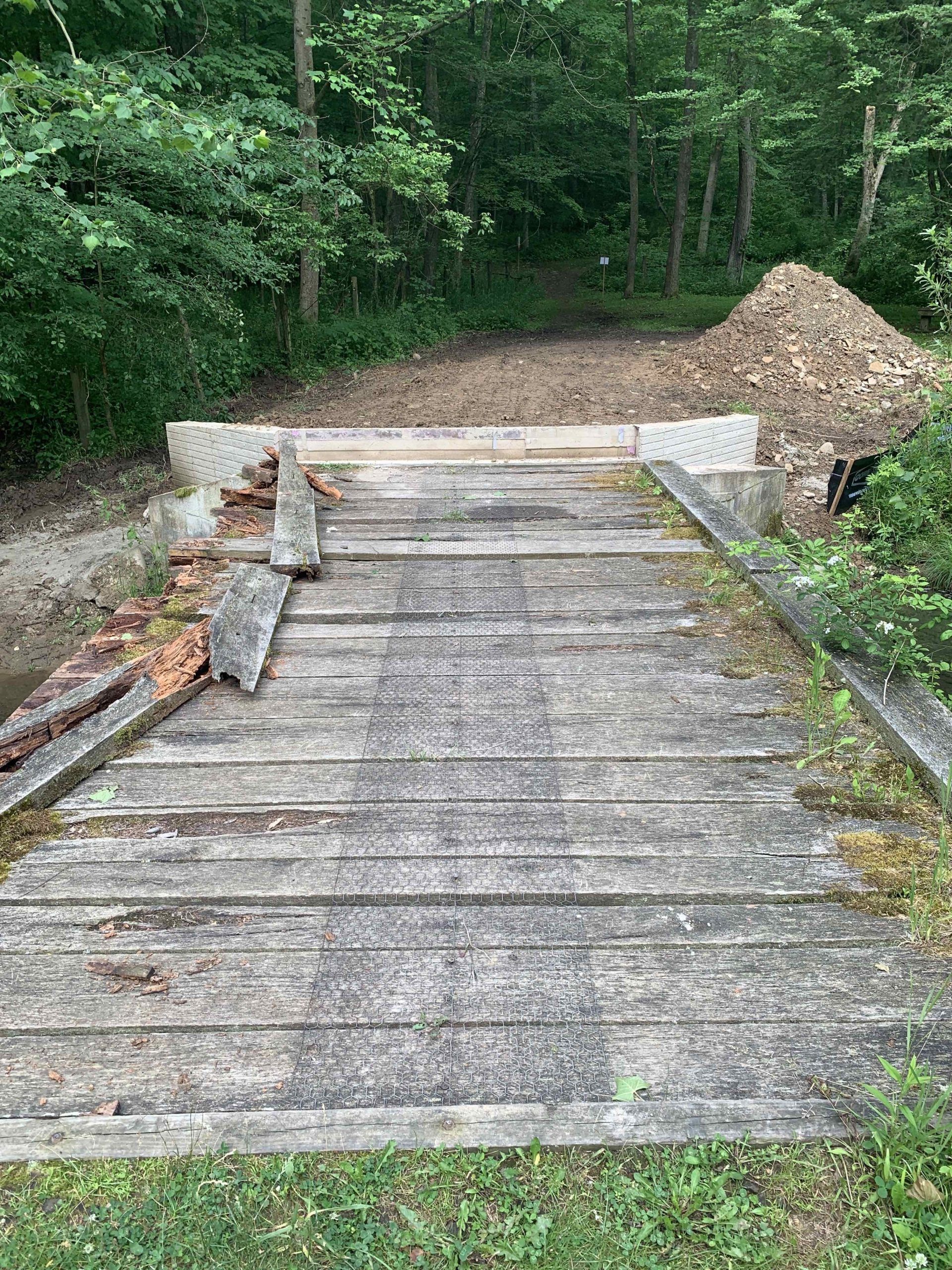 Damaged wooden bridge over a small stream in a wooded area. Pile of wood debris nearby.