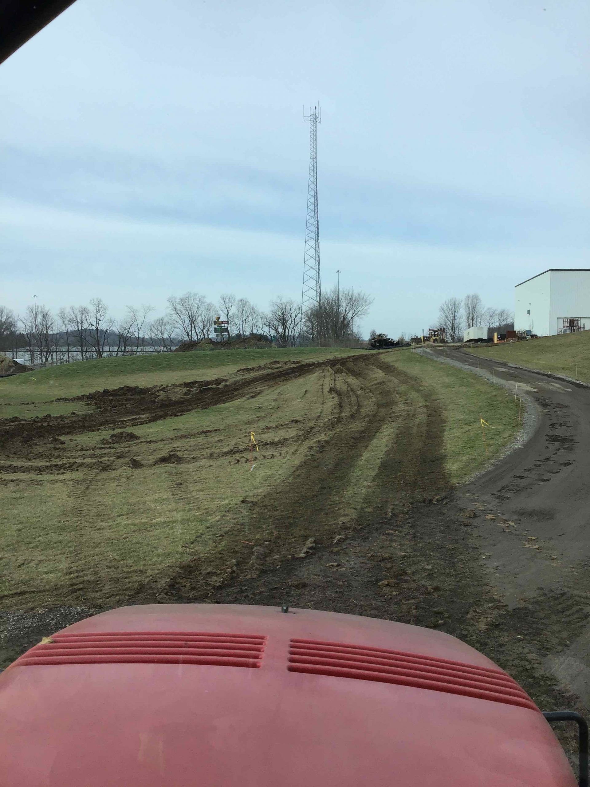 View from a red tractor cab, showing a muddy path and field with a radio tower in the distance.