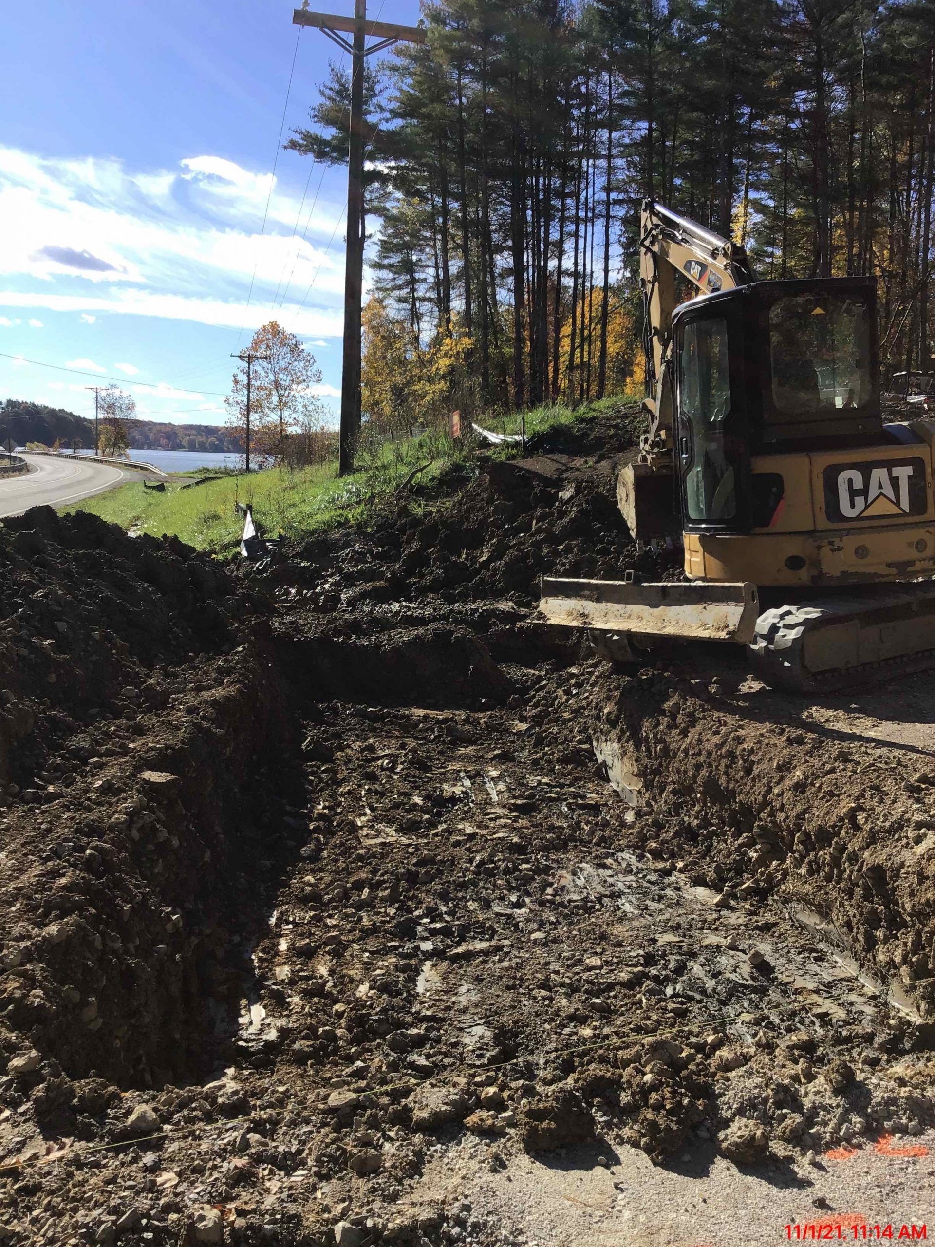 A yellow CAT excavator digging a trench on a roadside with a lake and trees in the background.