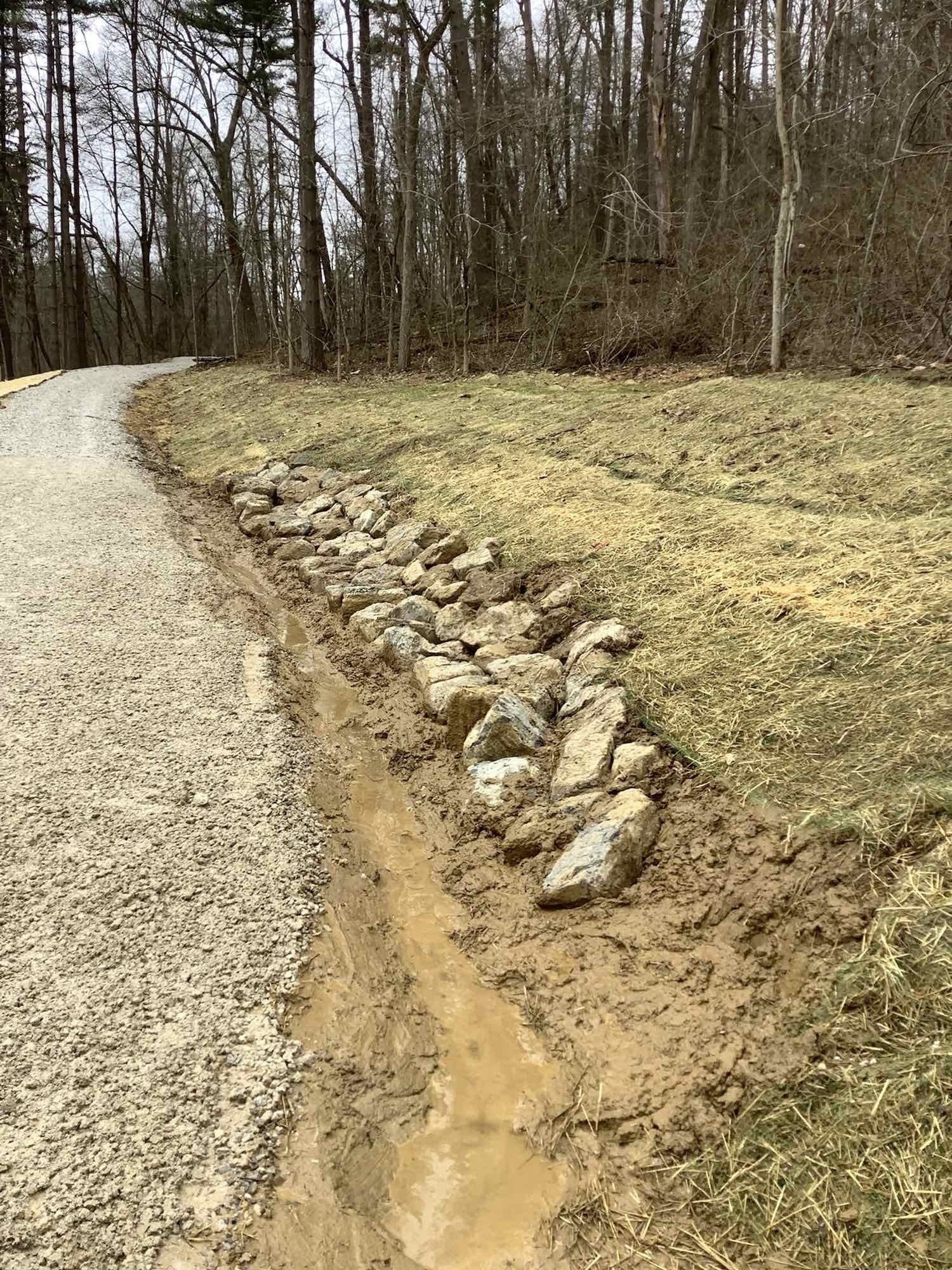 Gravel road with a rock-lined drainage ditch. Muddy, grass and trees in the background.