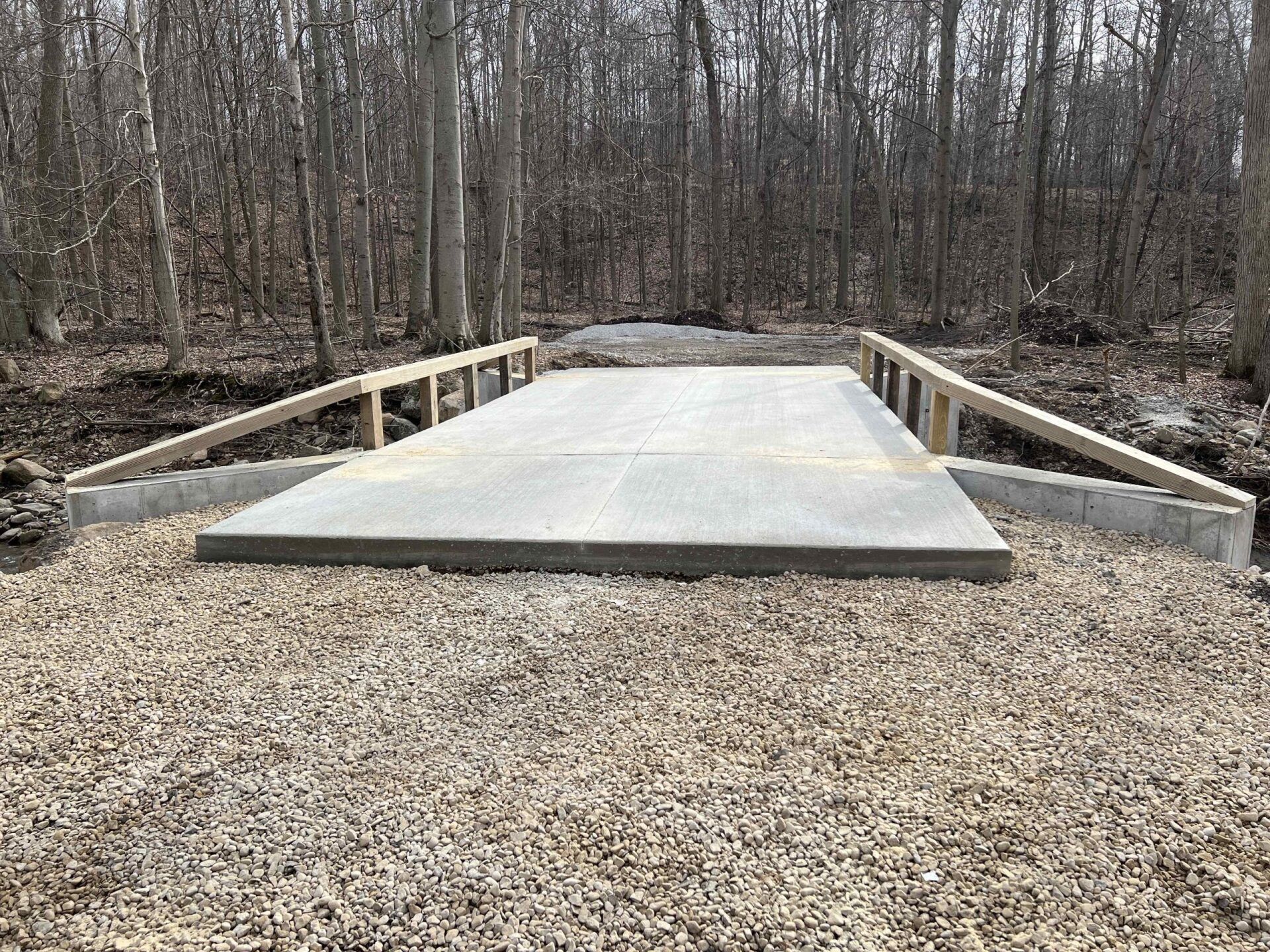 Concrete bridge with wood railings over gravel path in a wooded area.