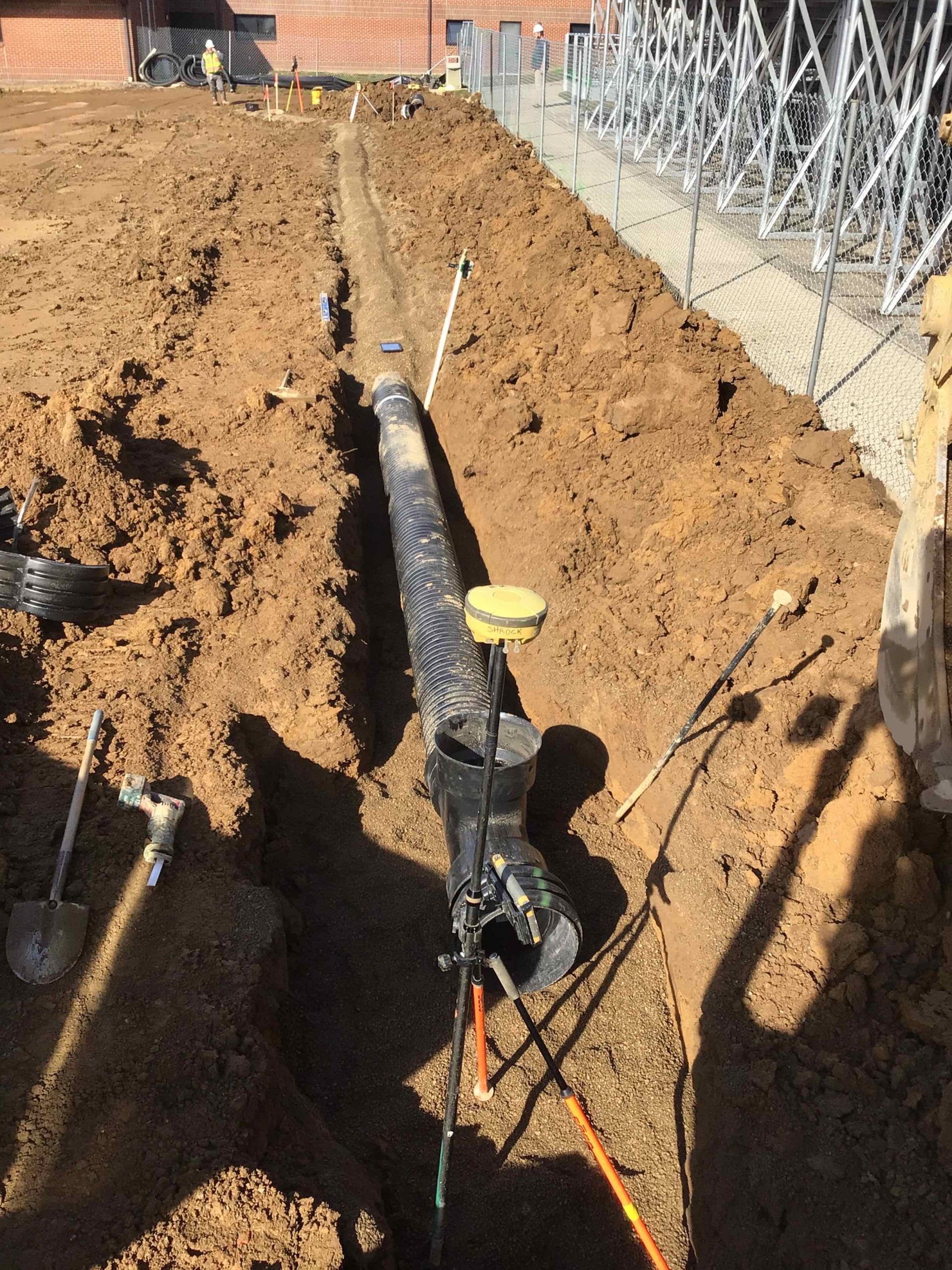 Trench in dirt with black pipe; GPS device attached. Construction site setting.