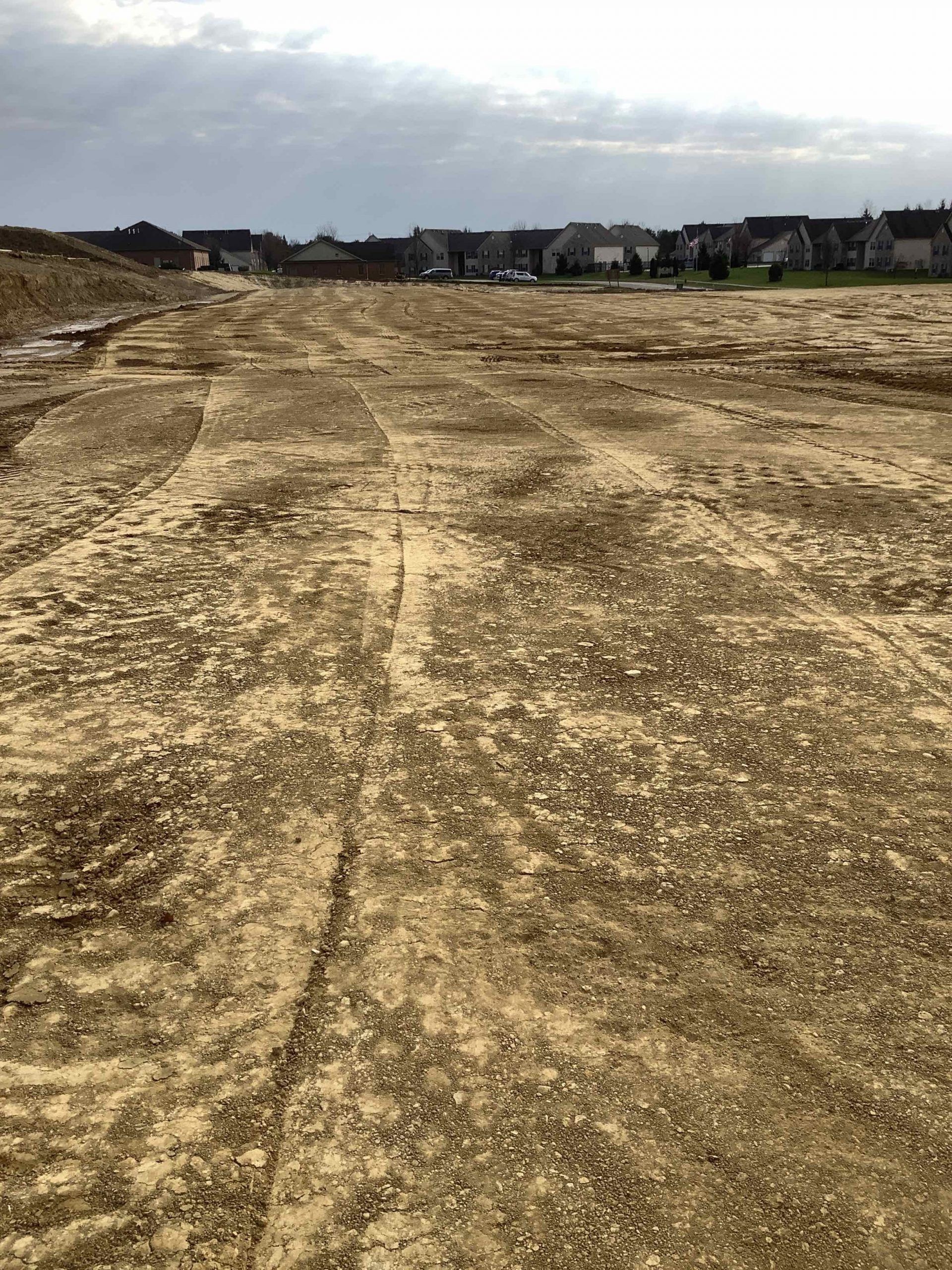 An empty, dirt construction site with tire tracks, houses in the distance under a cloudy sky.