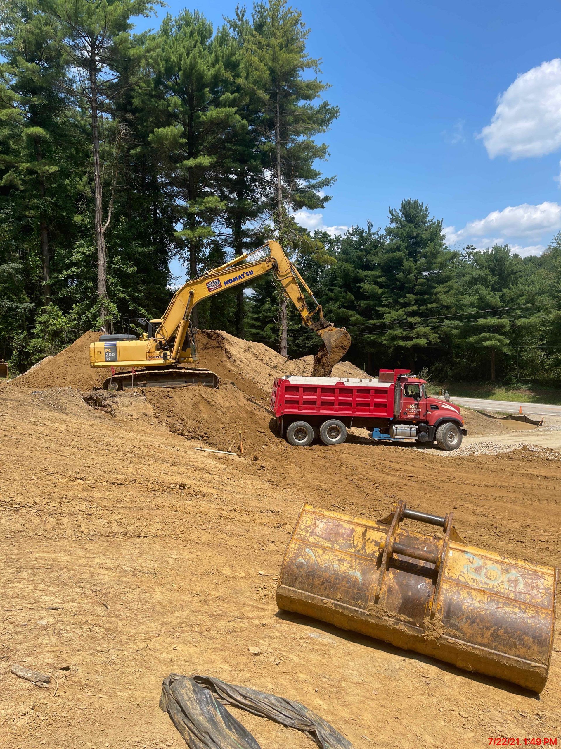 An excavator loading a red dump truck with dirt on a construction site, near trees under a blue sky.