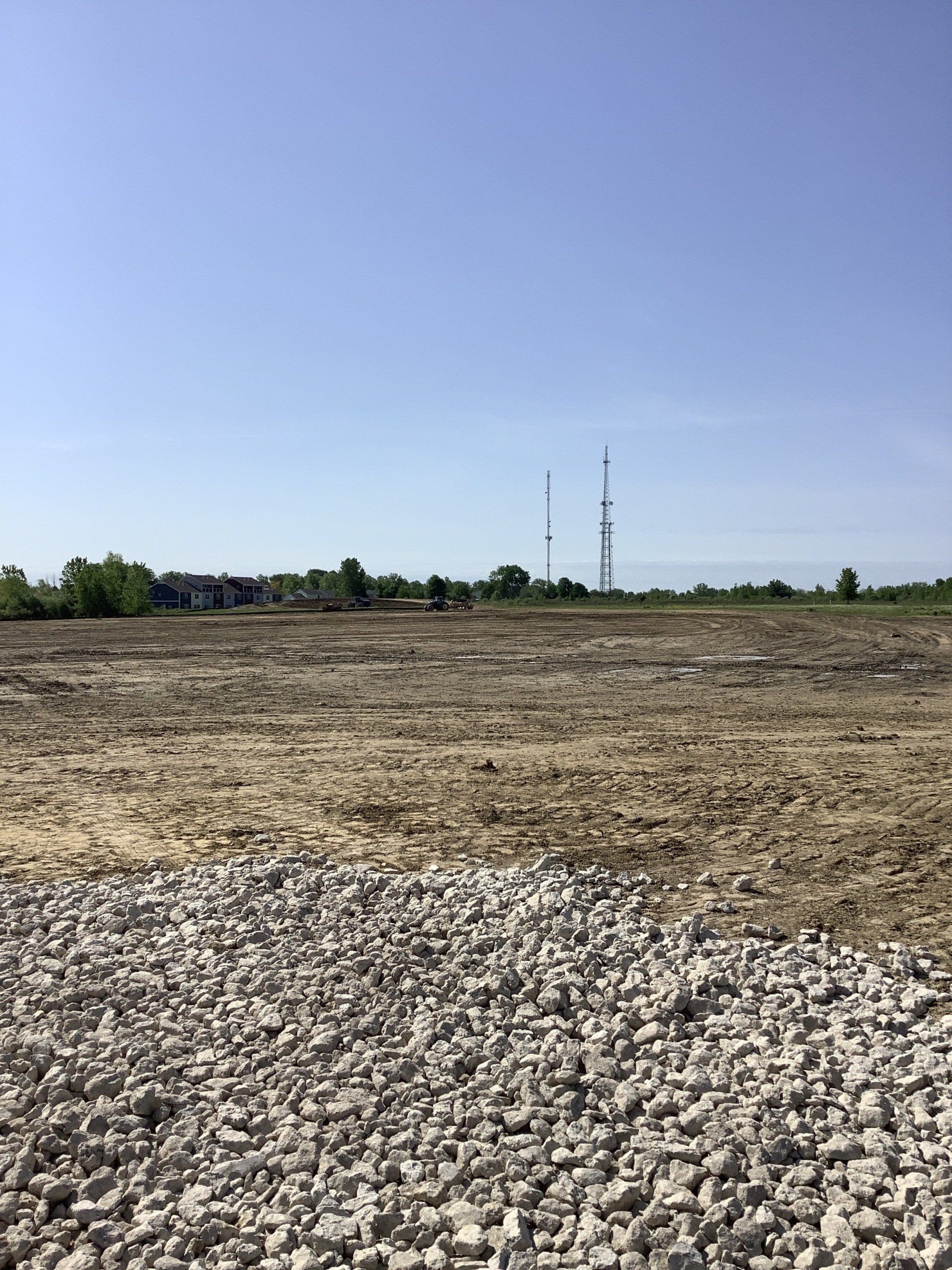 Cleared dirt lot with gravel in foreground, under a blue sky, and two tall towers in the distance.
