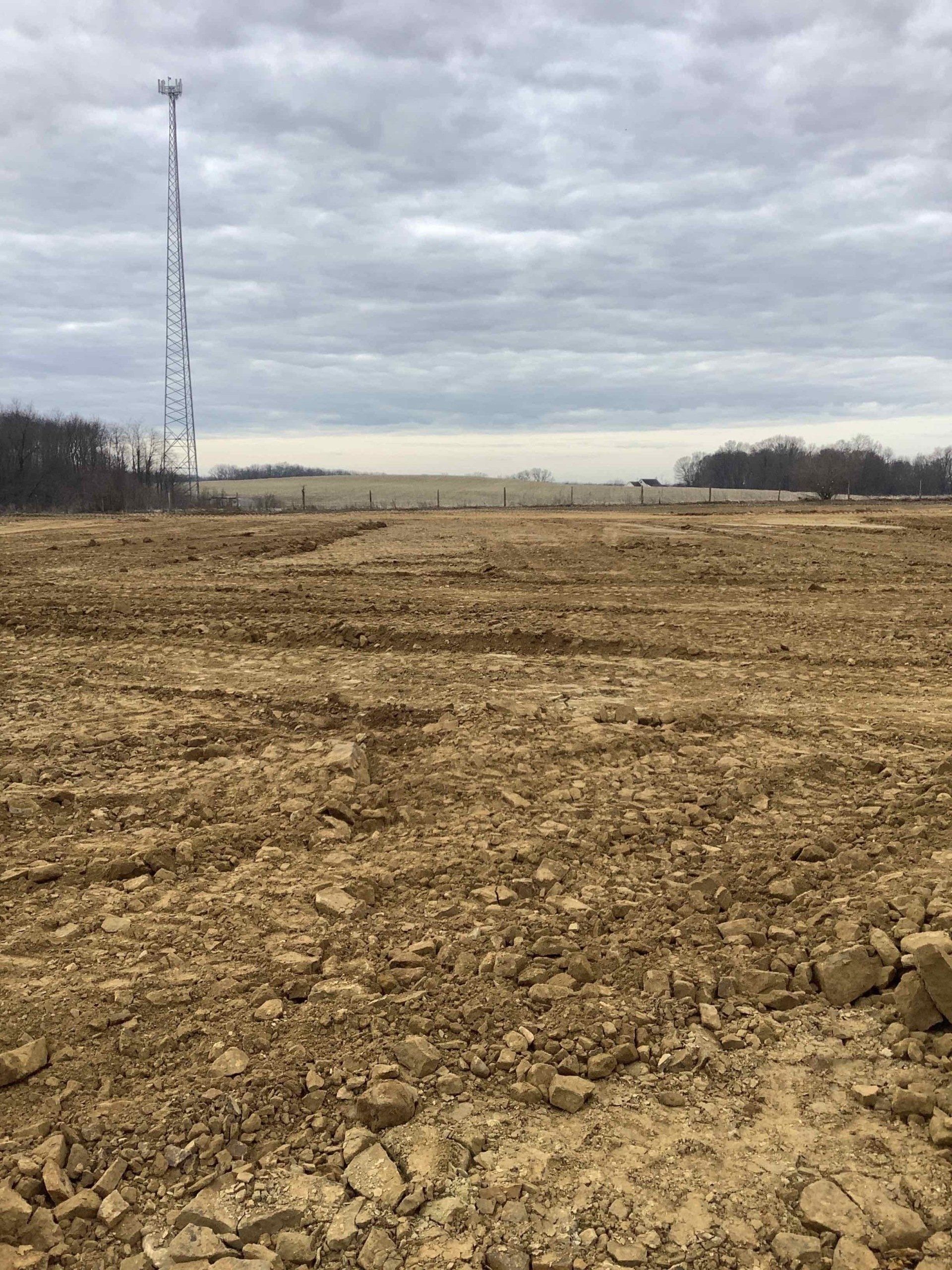 Brown, barren field with a cell tower in the distance under a cloudy sky.