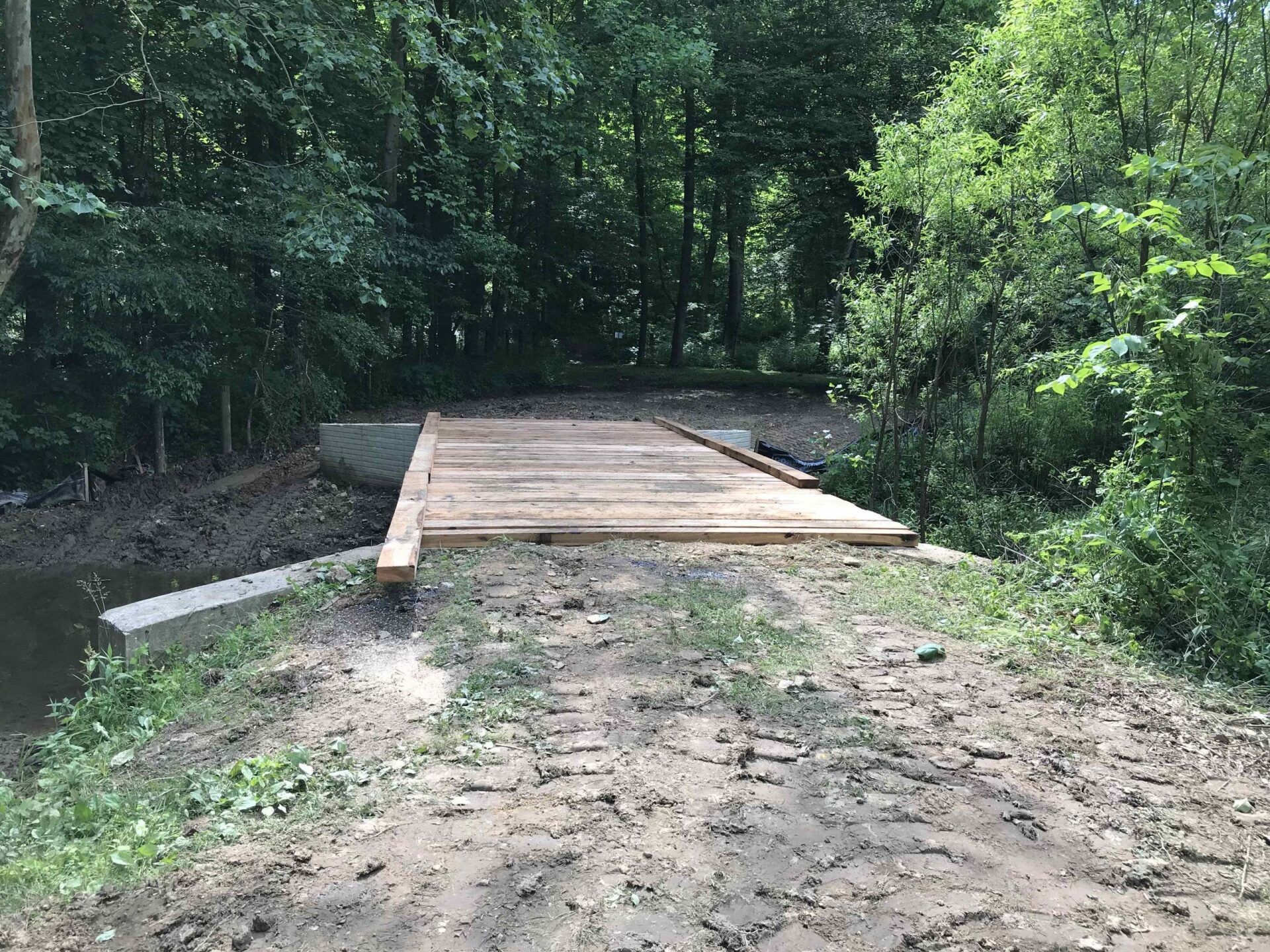 Wooden bridge over muddy terrain in a wooded area, supported by concrete structures.