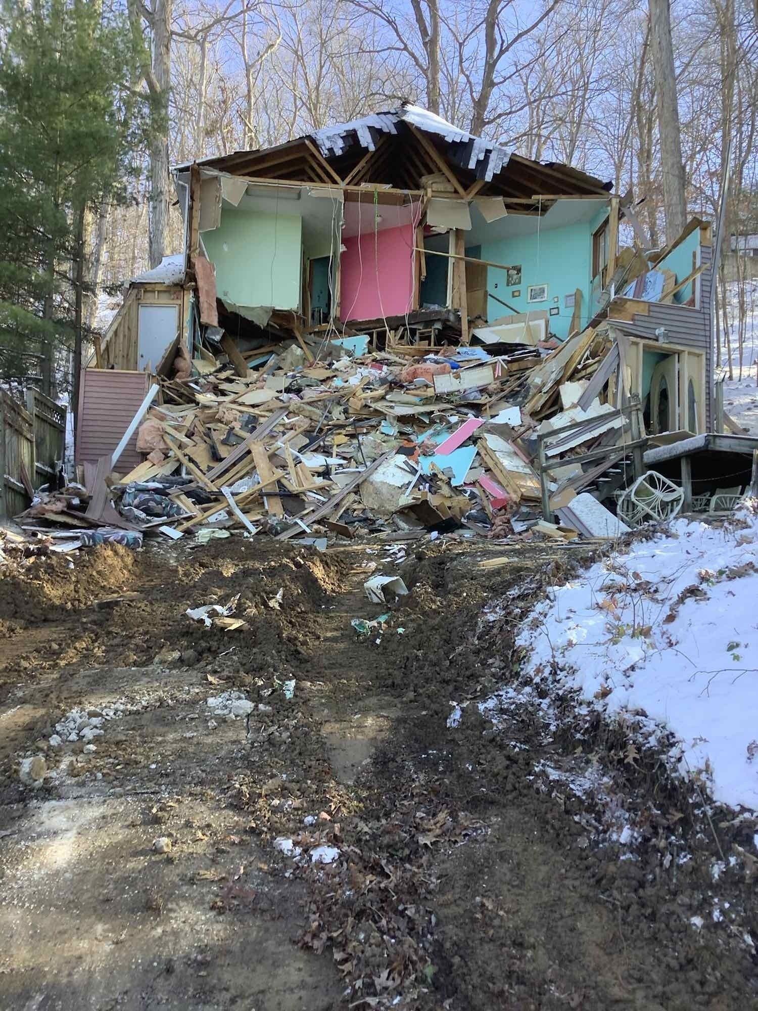Collapsed house; debris pile in front. Rooms display green, pink, and blue walls. Snowy, wooded setting.