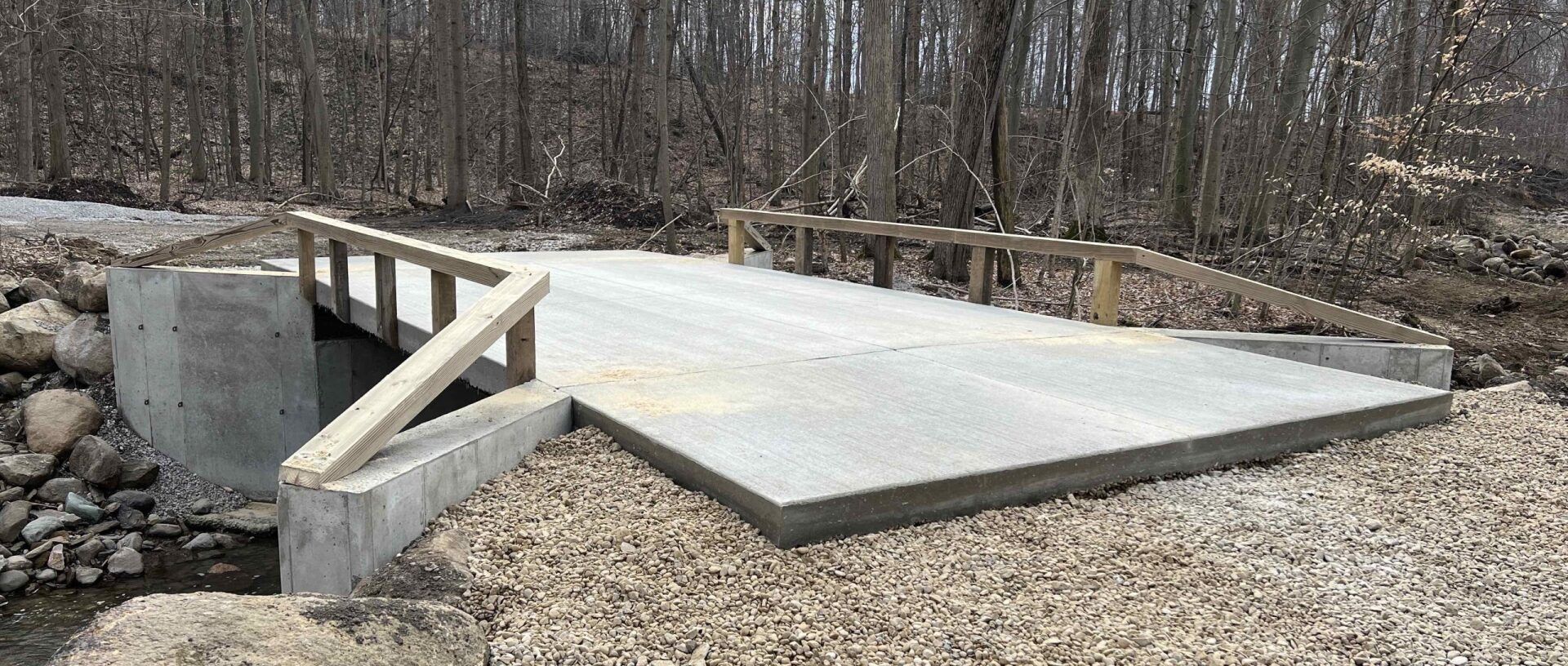 A concrete bridge with wooden railings over a rocky creek bed, surrounded by trees.