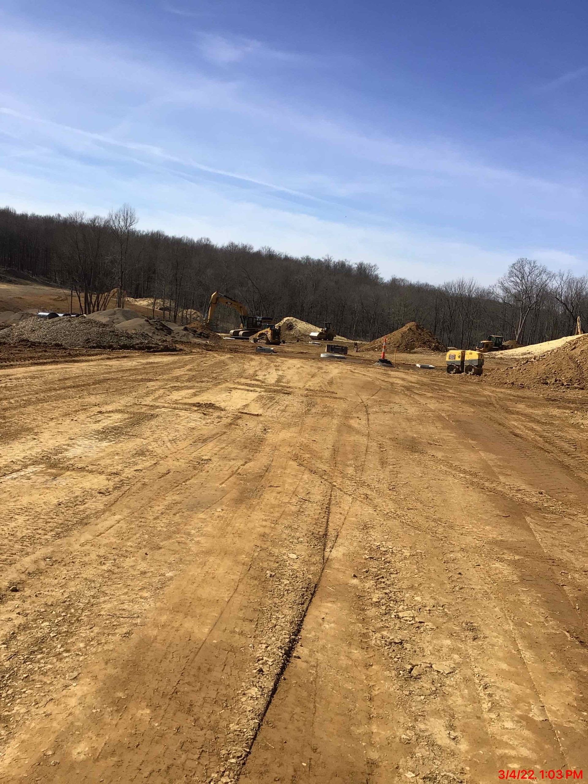 Dirt road leads to construction site with machinery, trees, and blue sky.