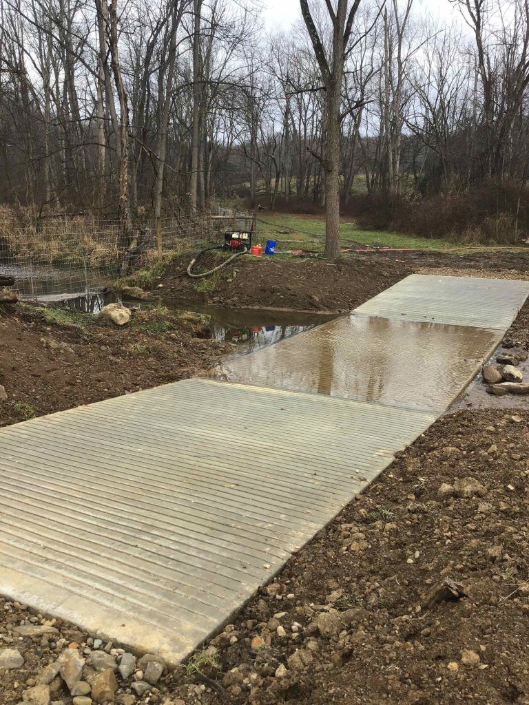 Concrete slabs forming a low-water bridge over a stream in a wooded area. The slabs have a textured surface.