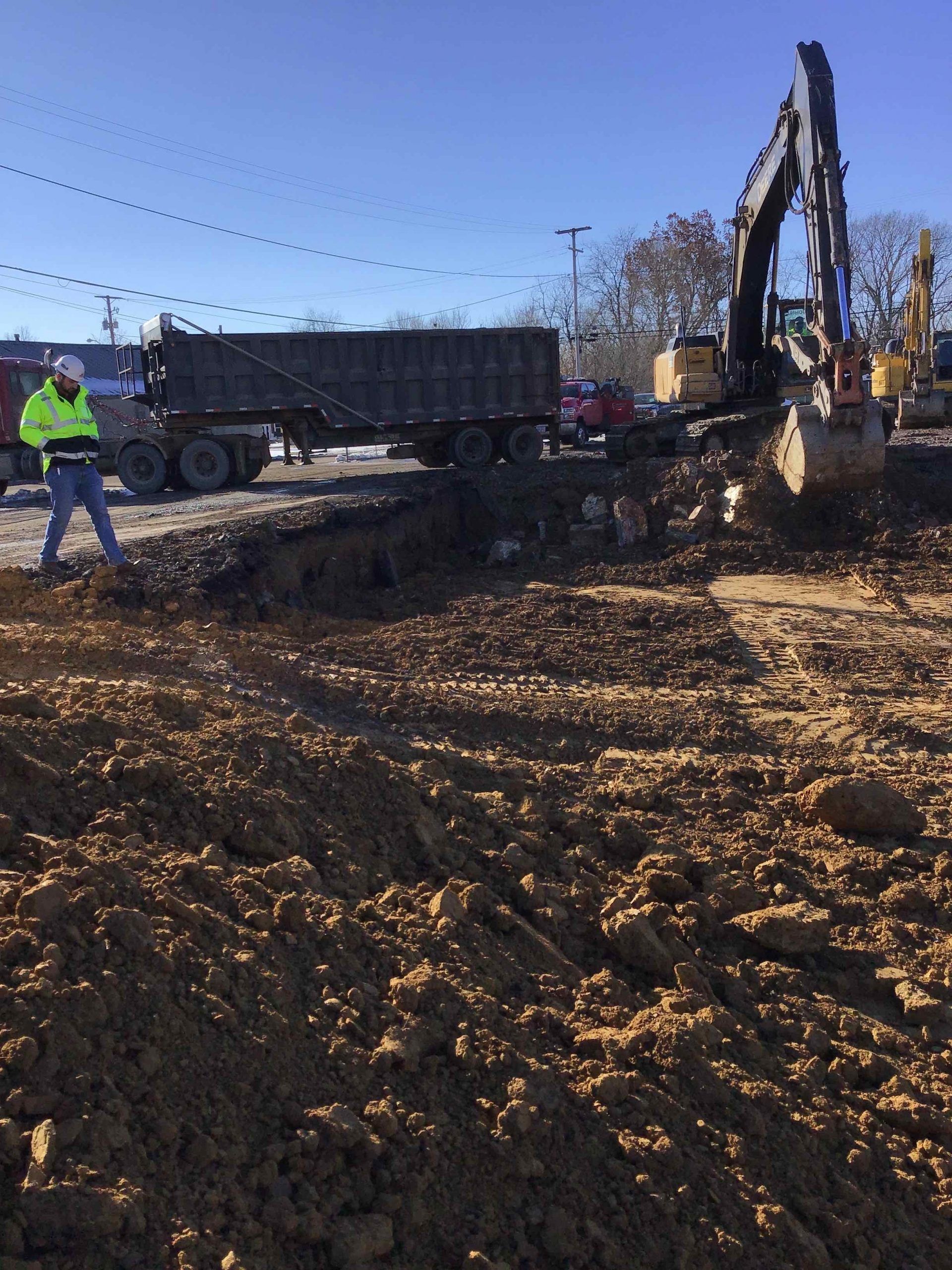 Construction site: excavator loading dirt into a dump truck, worker in neon vest observes.