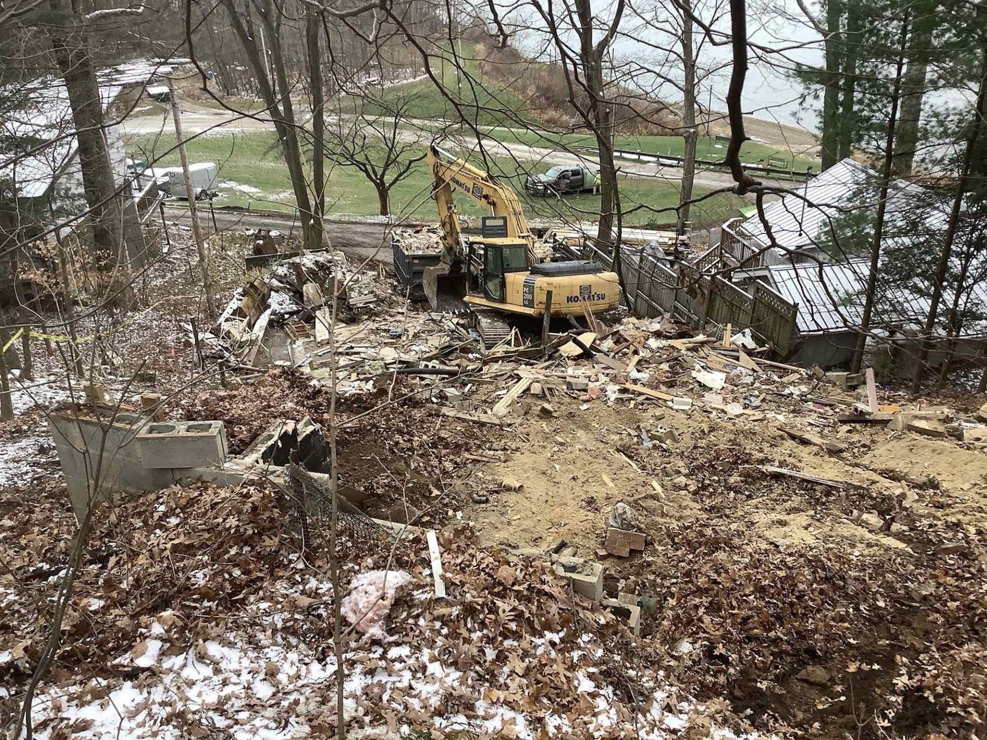 Yellow excavator demolishing debris in a wooded area, with snowy ground and bare trees.