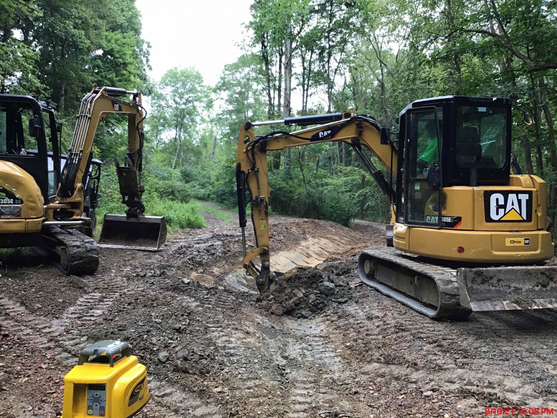 Three yellow CAT mini excavators working on a muddy site surrounded by trees.