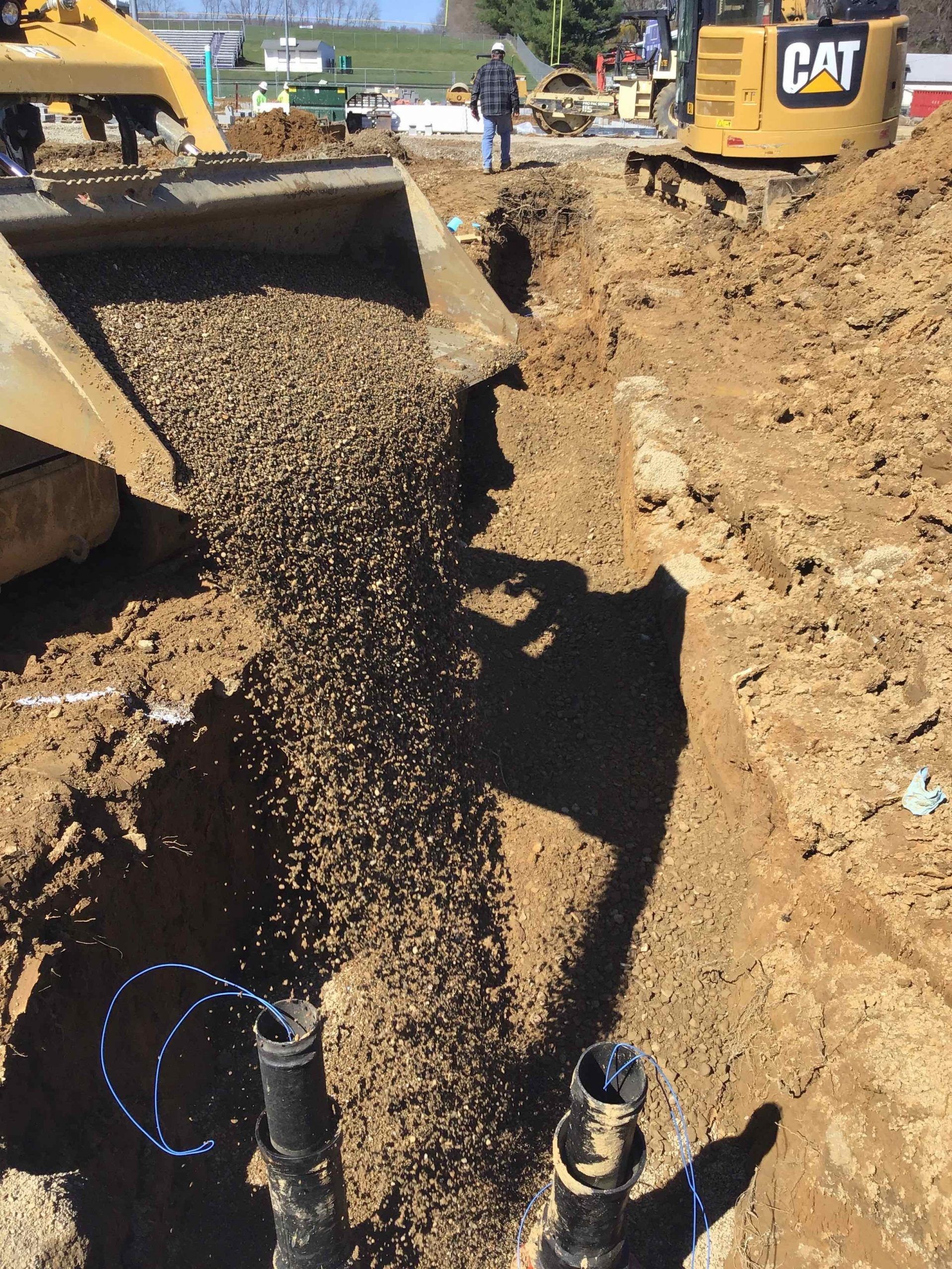 A backhoe dumping gravel into a trench containing two black pipes at a construction site. Construction worker in background. 
