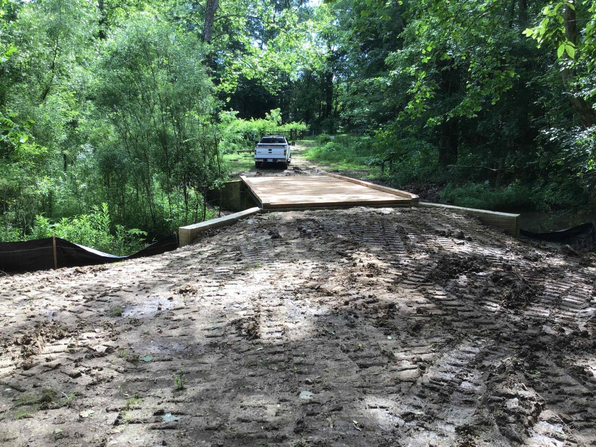 Muddy dirt road leads to a wooden bridge with a truck parked on it, surrounded by trees.