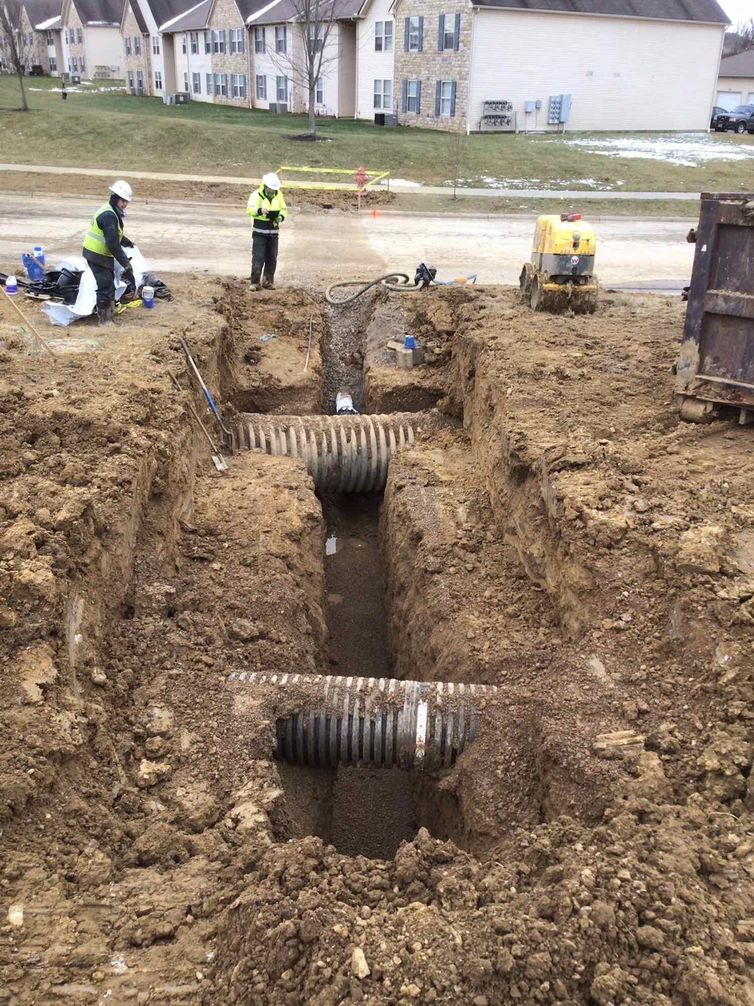 Workers in safety vests in a construction site trench, installing corrugated pipes near residential buildings.
