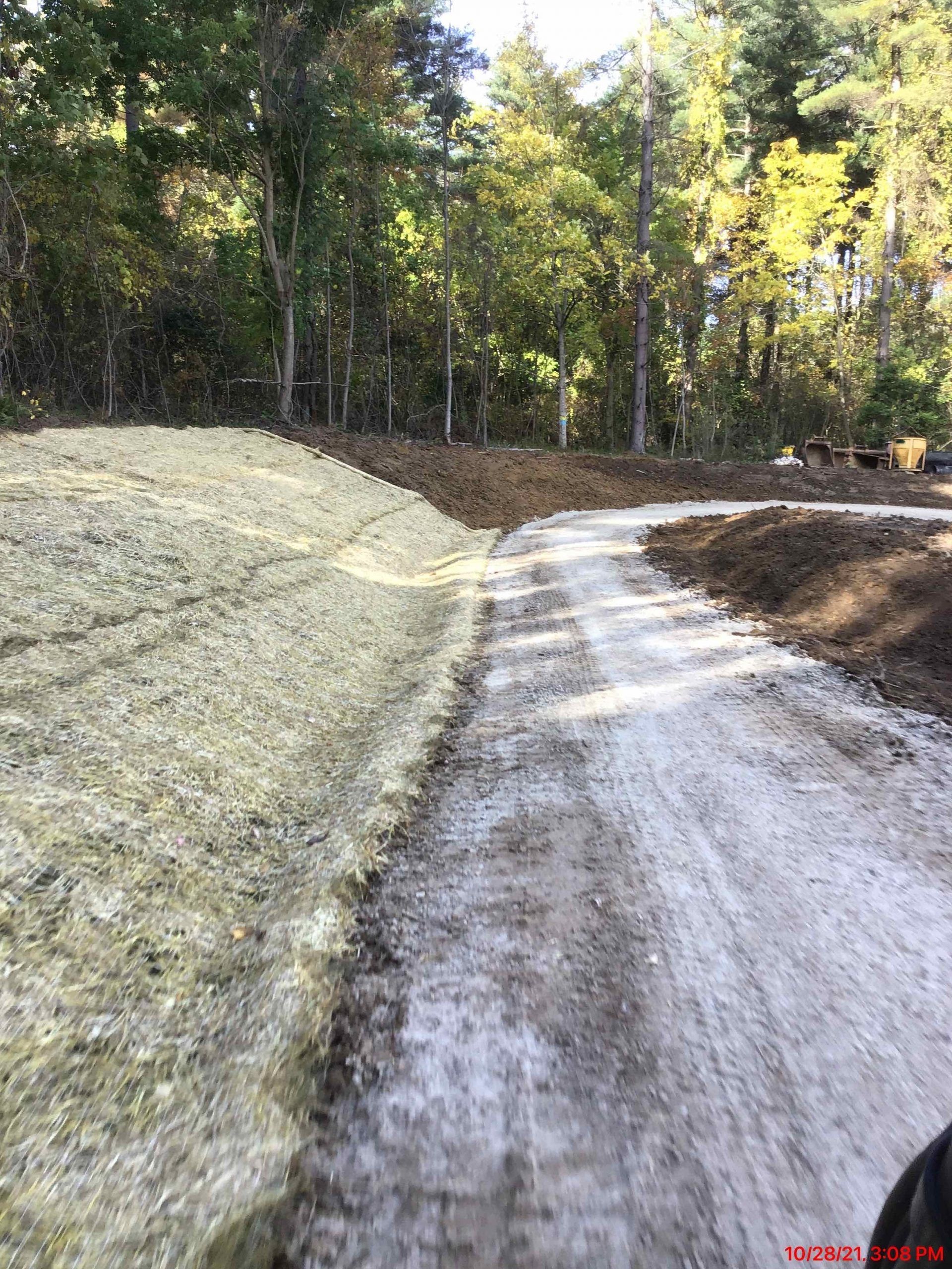Dirt road curves through a wooded area; a pile of tan gravel on the left.