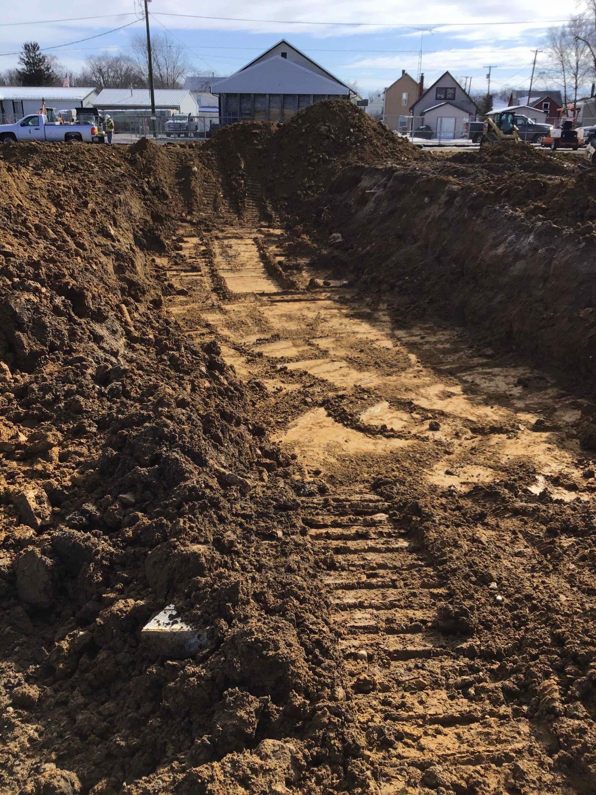 Trench in dirt with tractor tracks, building and vehicles in background.