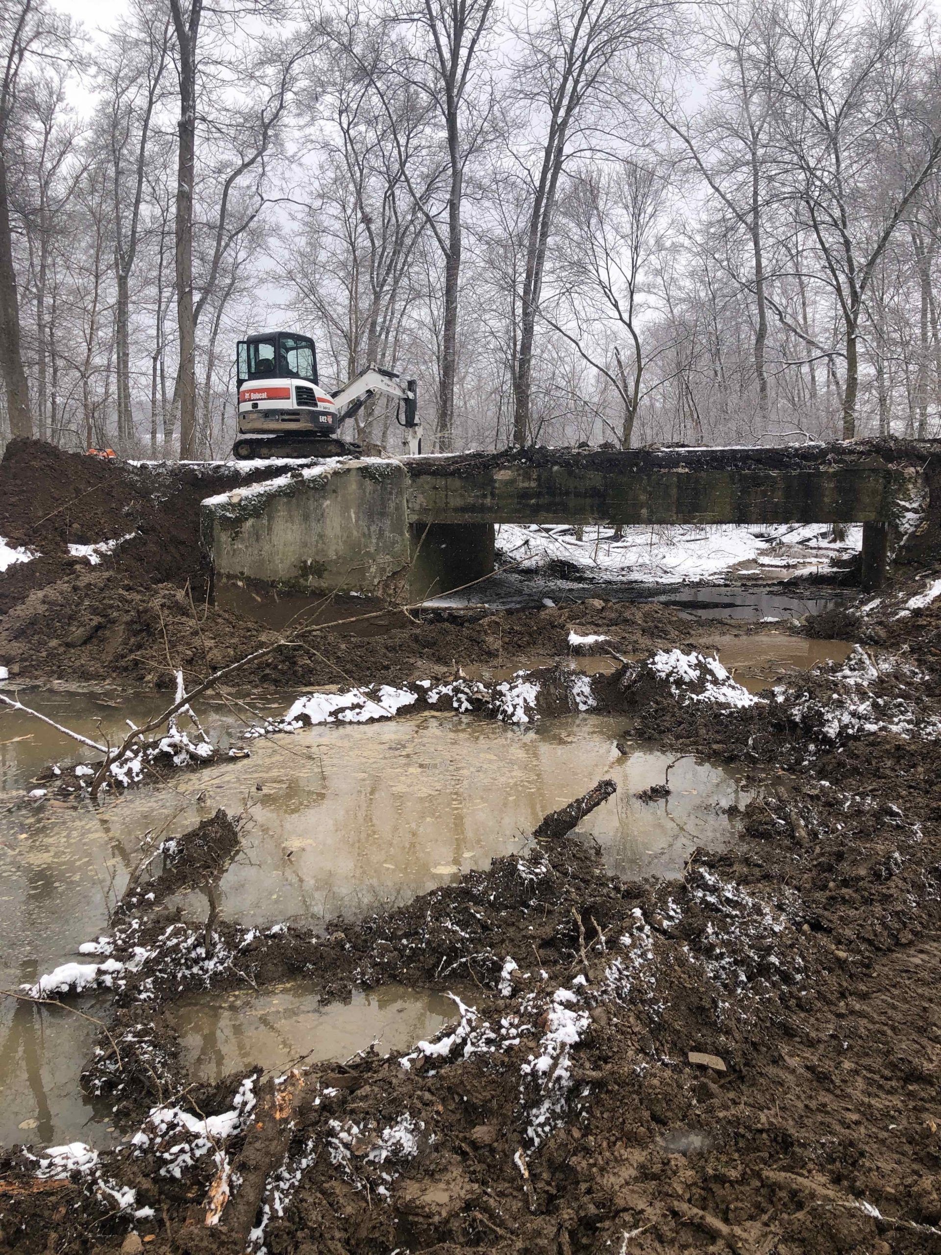 Construction site with muddy ground, small excavator, and concrete bridge in a snowy forest.