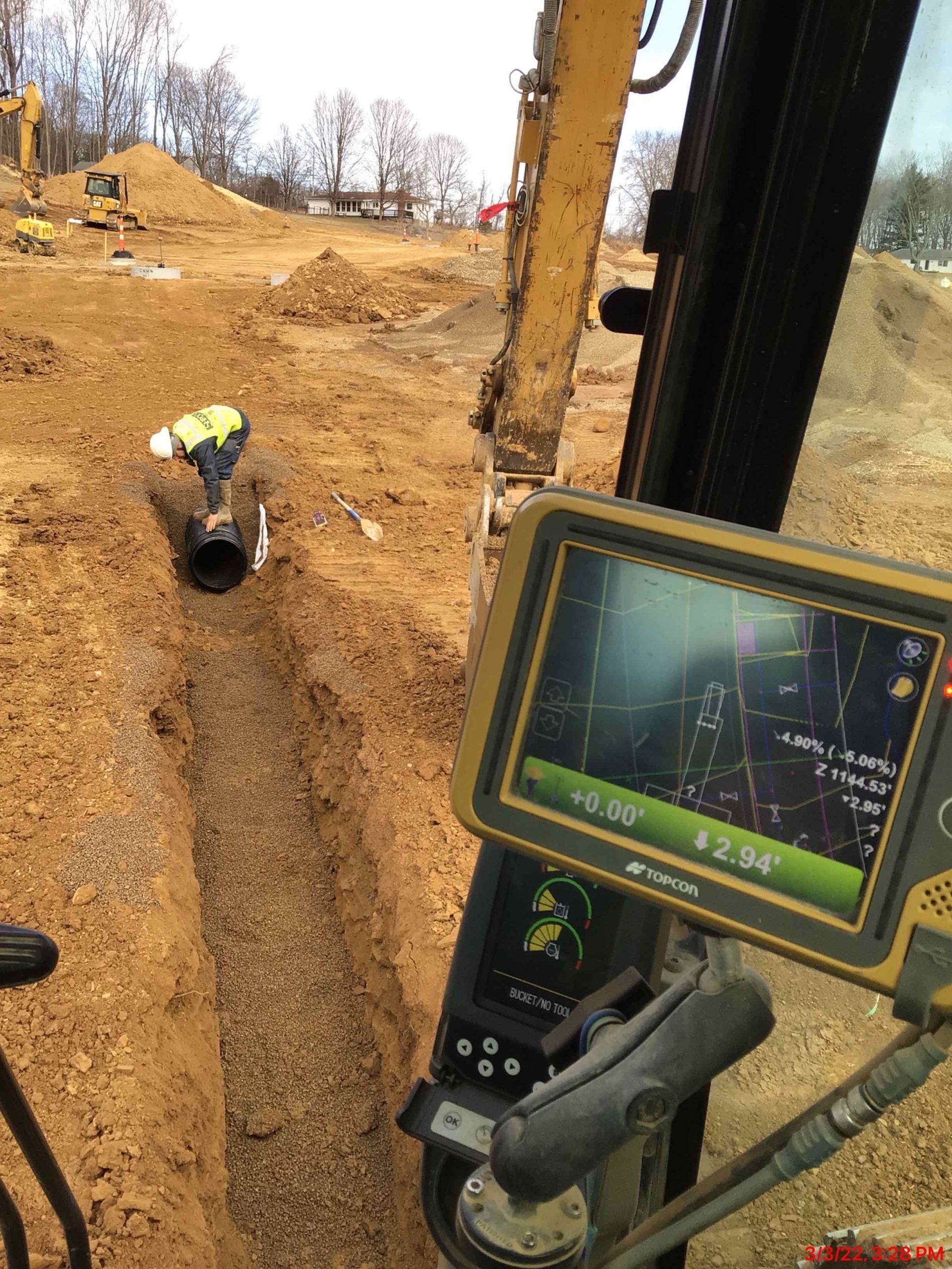 An excavator's monitor displaying a map, with a worker in a trench. Construction site, brown soil.