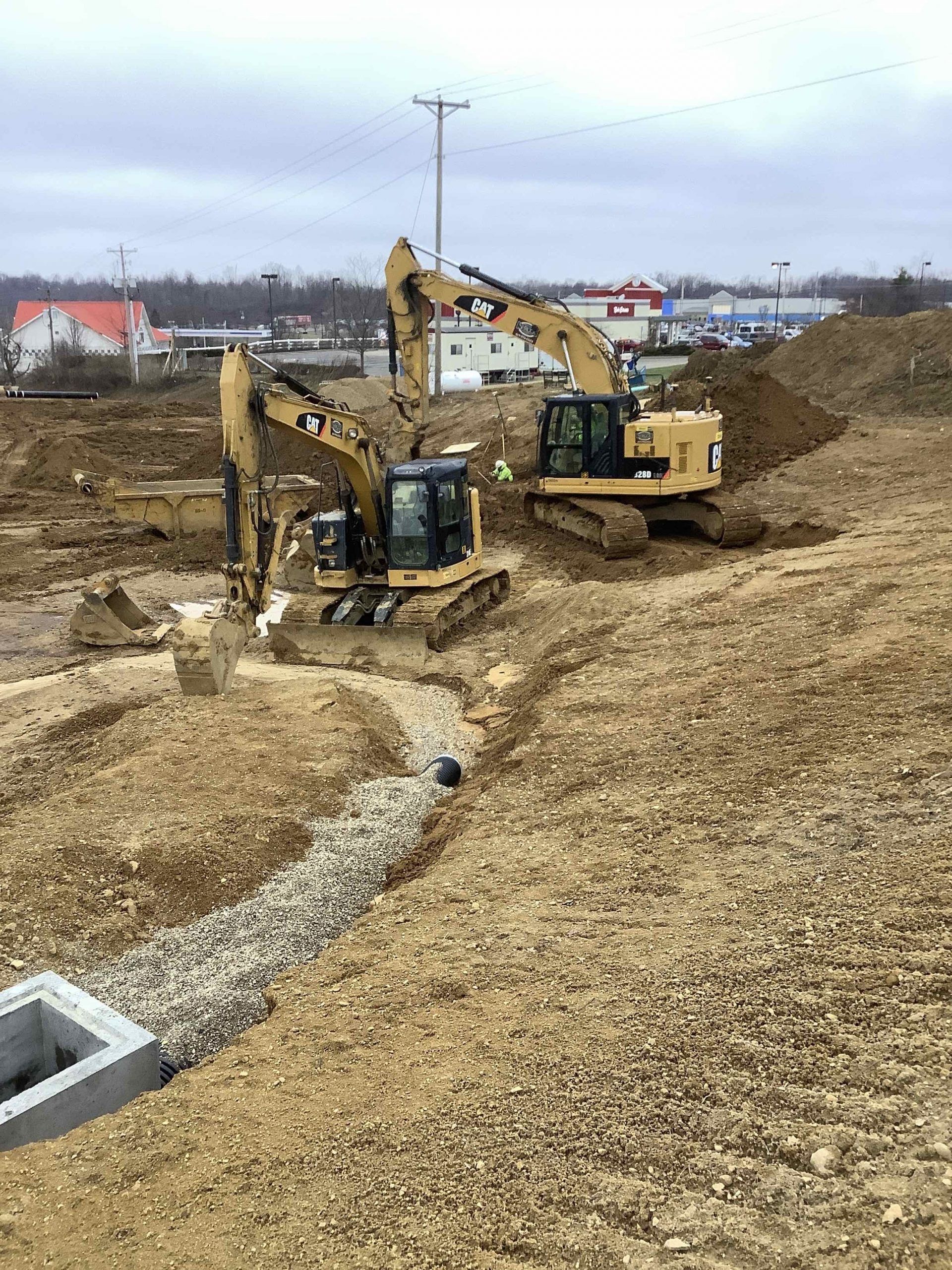 Two yellow excavators working on a construction site with a stone-lined drainage area and a concrete structure.