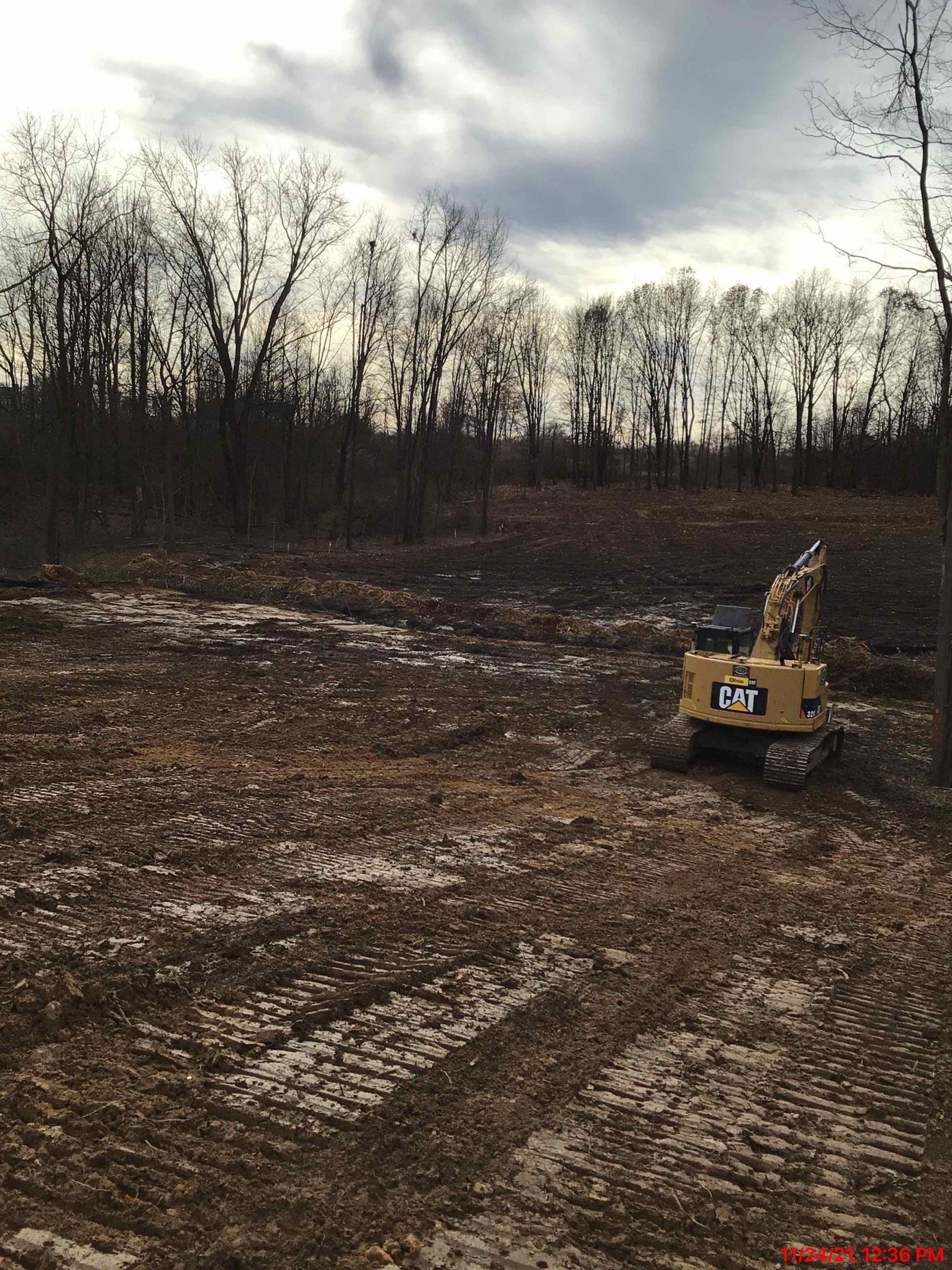 Yellow CAT excavator in a muddy field near bare trees under a cloudy sky.