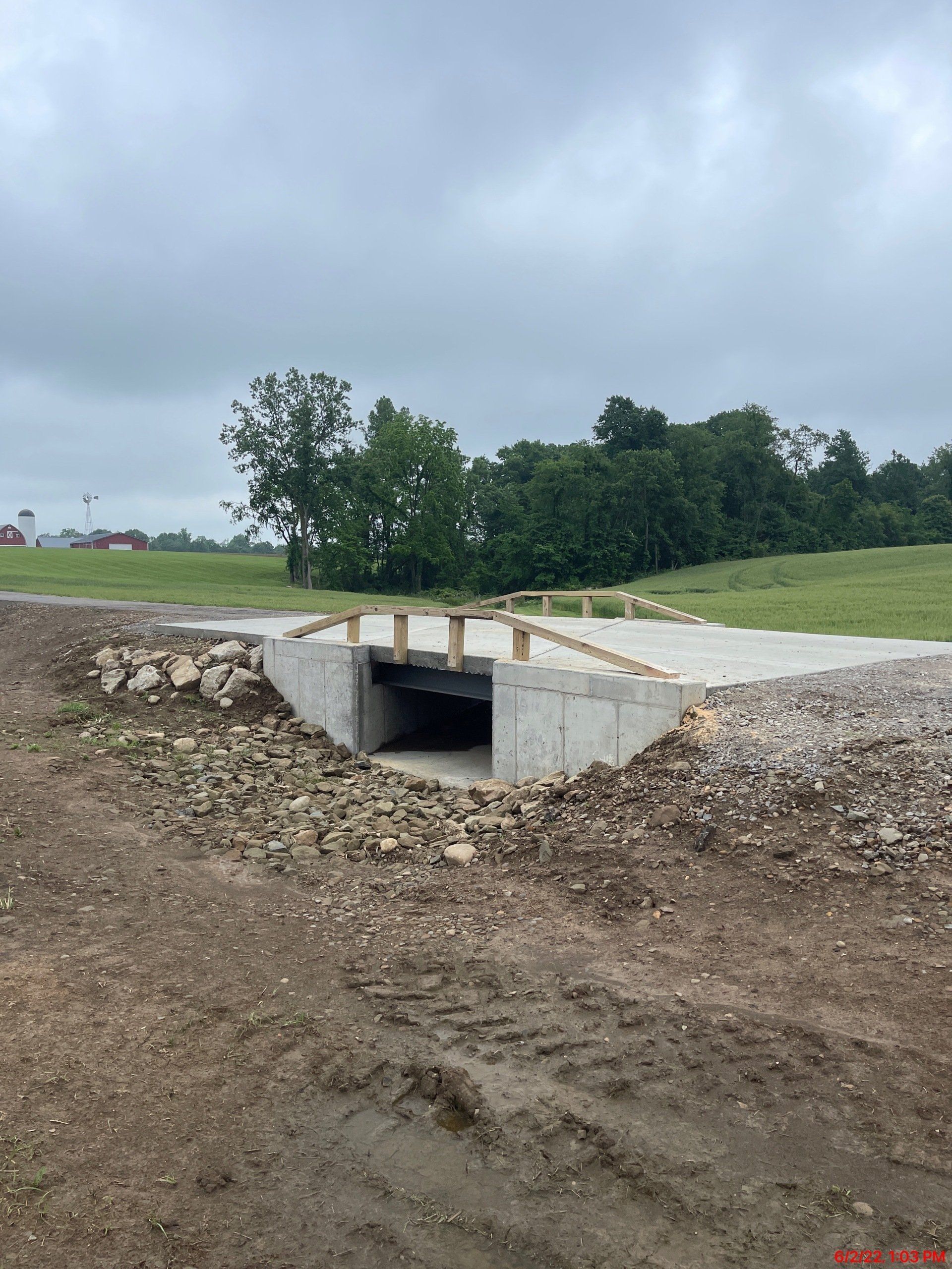 Concrete bridge over a small drainage ditch, gravel pathways on both sides, and grassy field beyond.