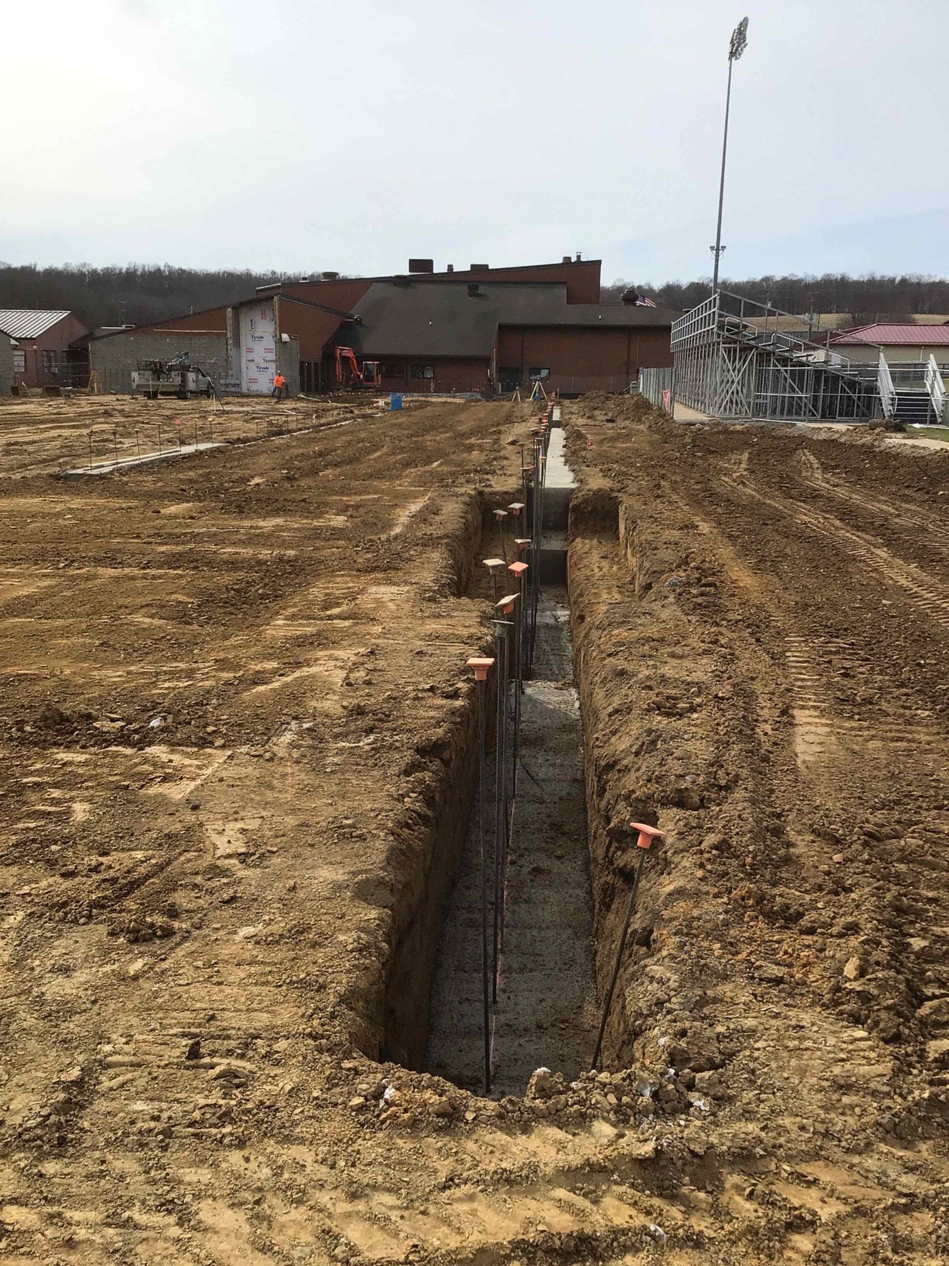 Trench dug in dirt, with concrete foundation being laid. Construction site with buildings in background.