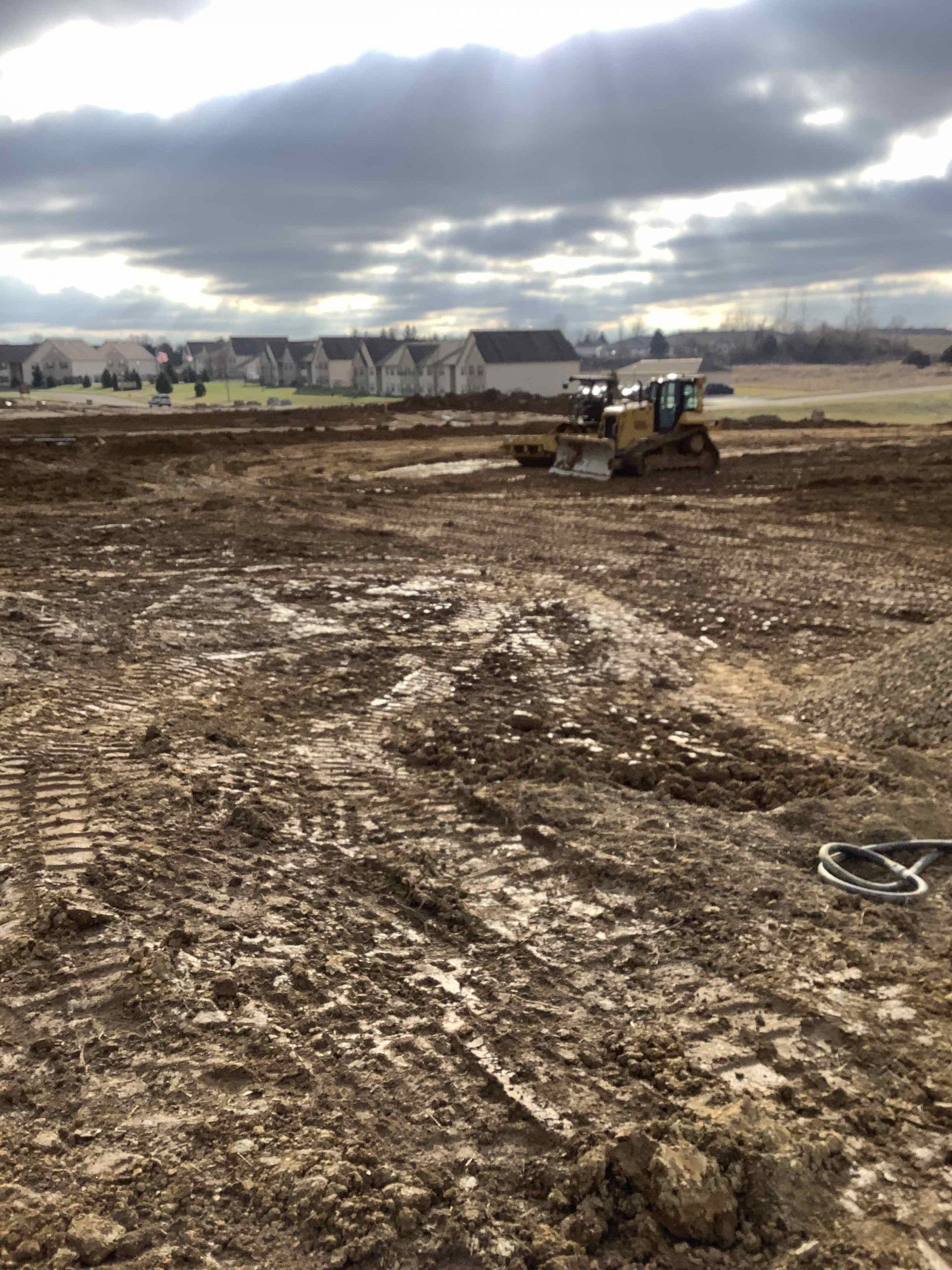Construction site with bulldozer flattening muddy ground; houses in the background under a cloudy sky.