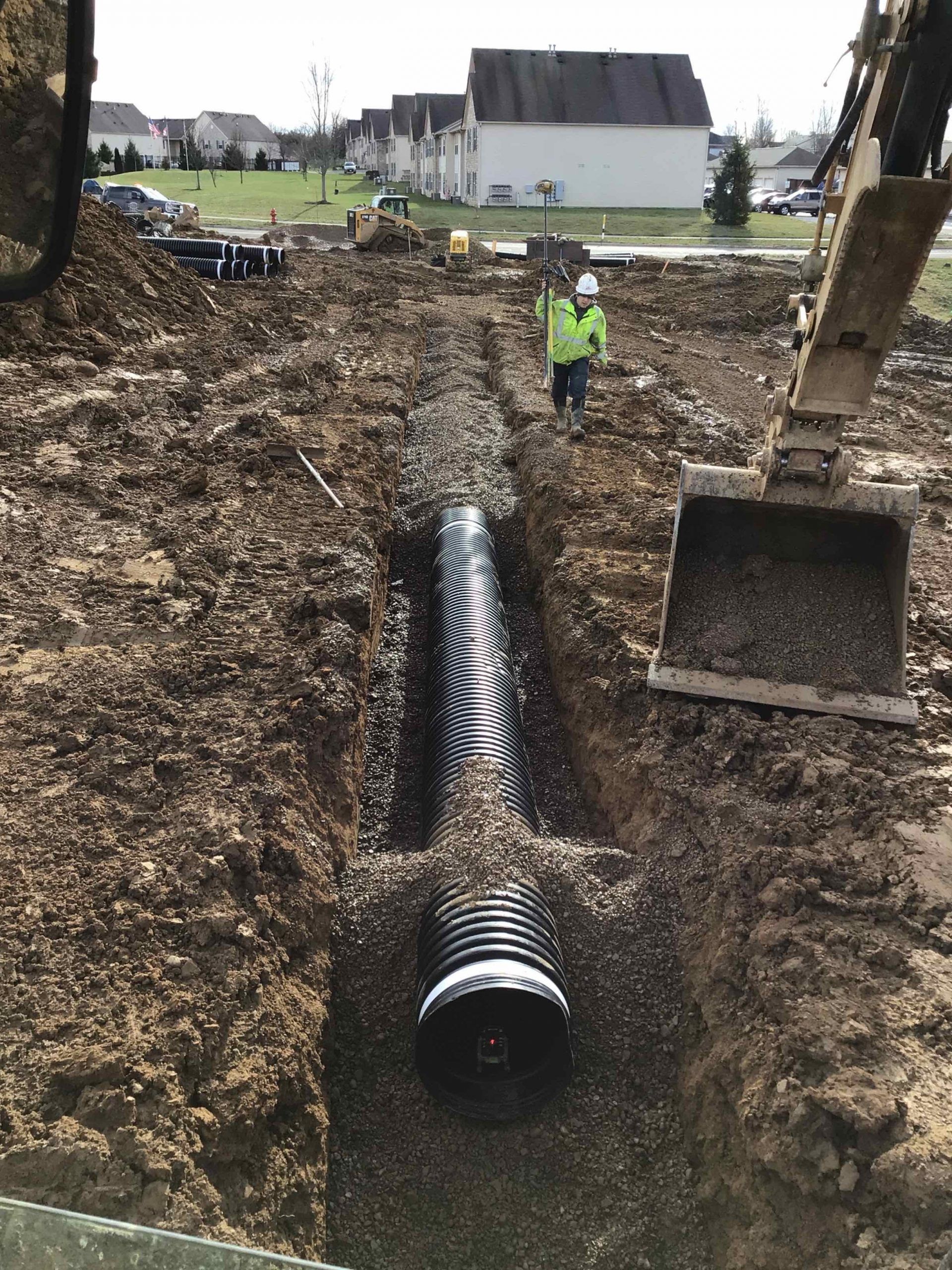 Construction site with a corrugated pipe in a trench, excavator, worker in reflective vest.