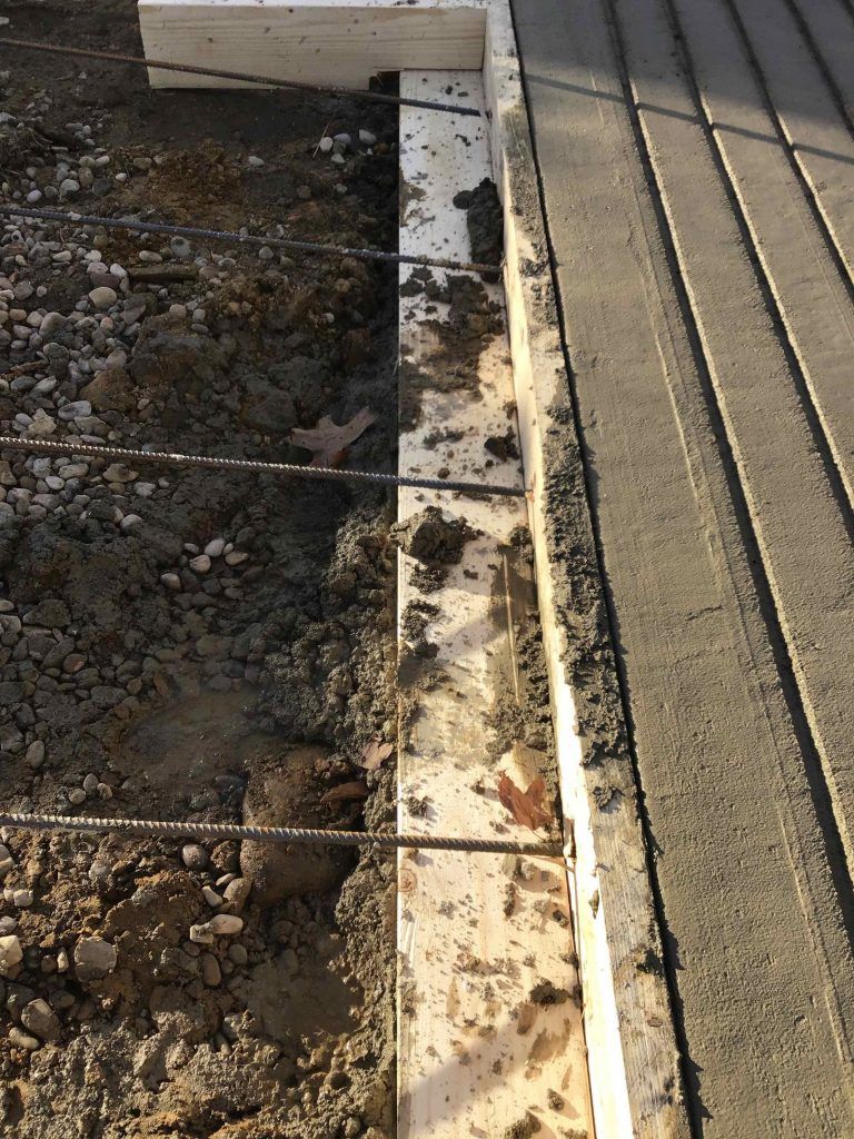 Concrete construction: Formwork with rebar next to poured concrete sidewalk. Soil and gravel visible.