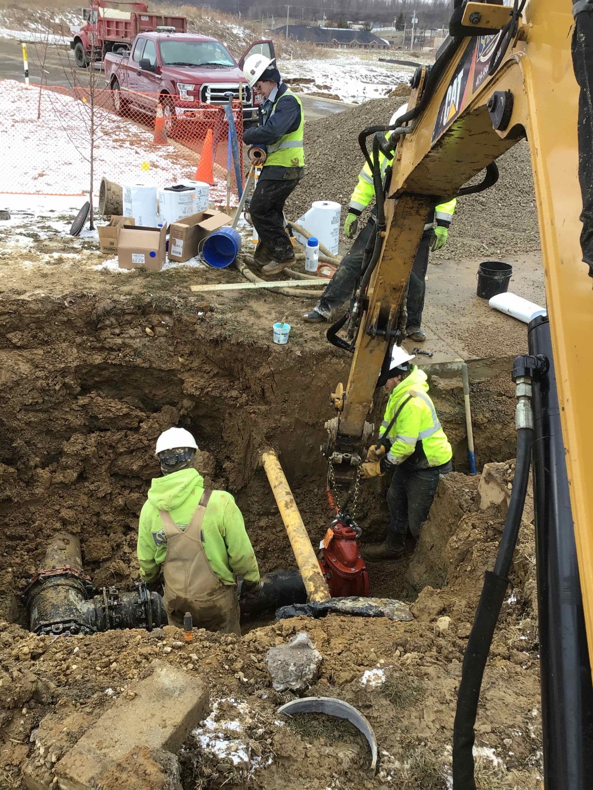 Workers in safety vests repair a pipe in a deep trench, using an excavator. Red truck and supplies are in background.