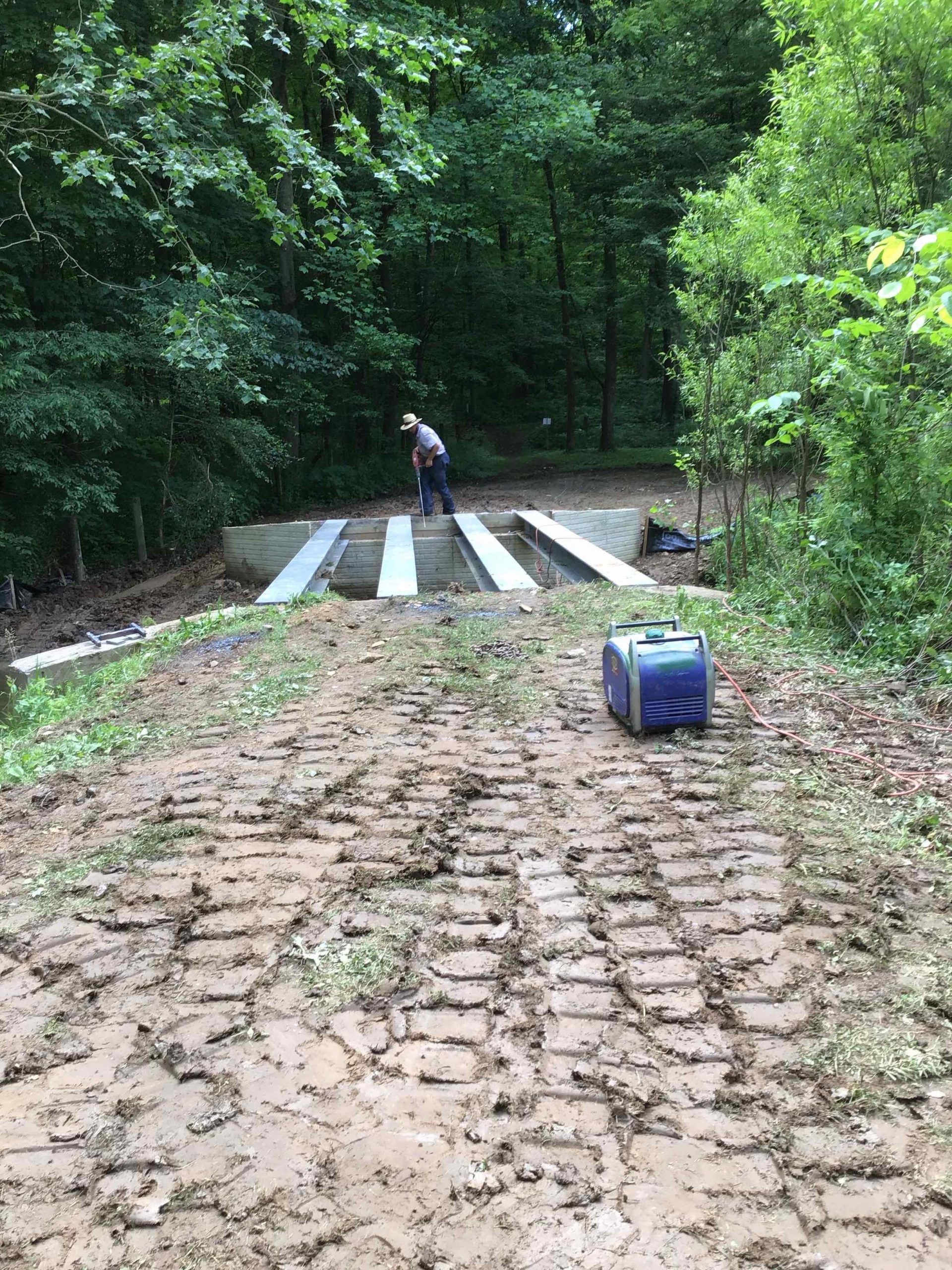 Muddy road leading to a concrete structure in a forest, with a person working on top and a blue container nearby.