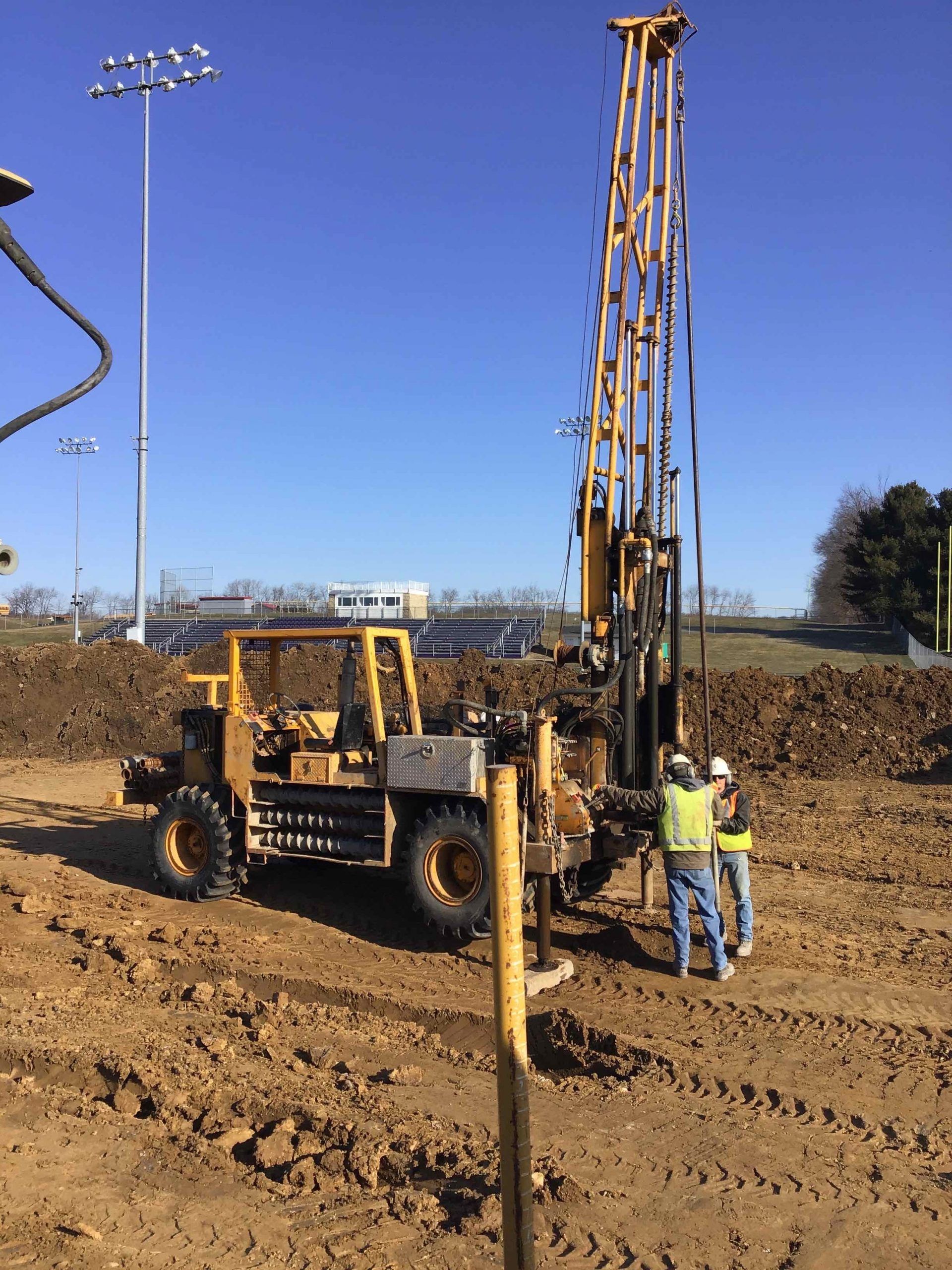 Yellow drilling rig on a construction site; two workers nearby. Clear, blue sky.