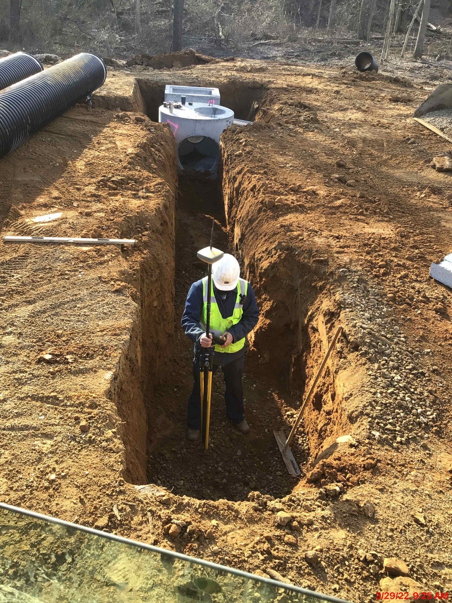 Construction worker in trench, installing drainage pipe. Yellow vest, hard hat, dirt. Outdoors.