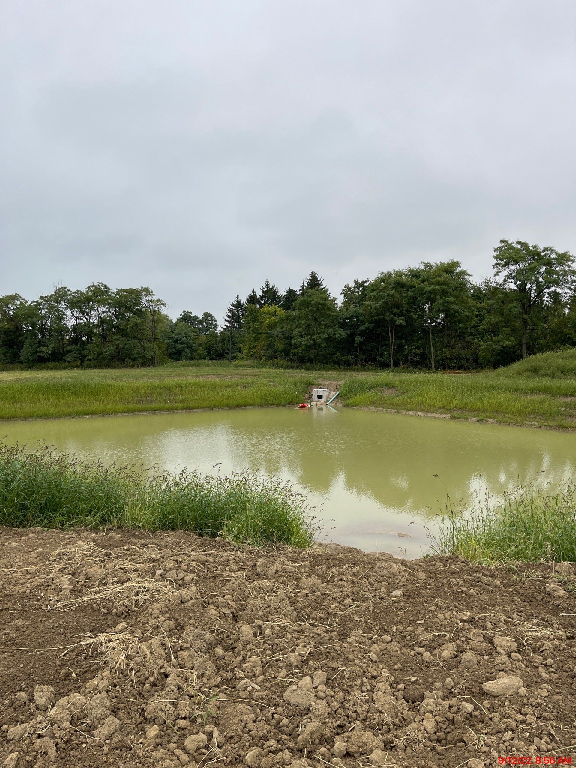 A muddy pond with a small concrete outlet, surrounded by dirt and green vegetation, under a cloudy sky.