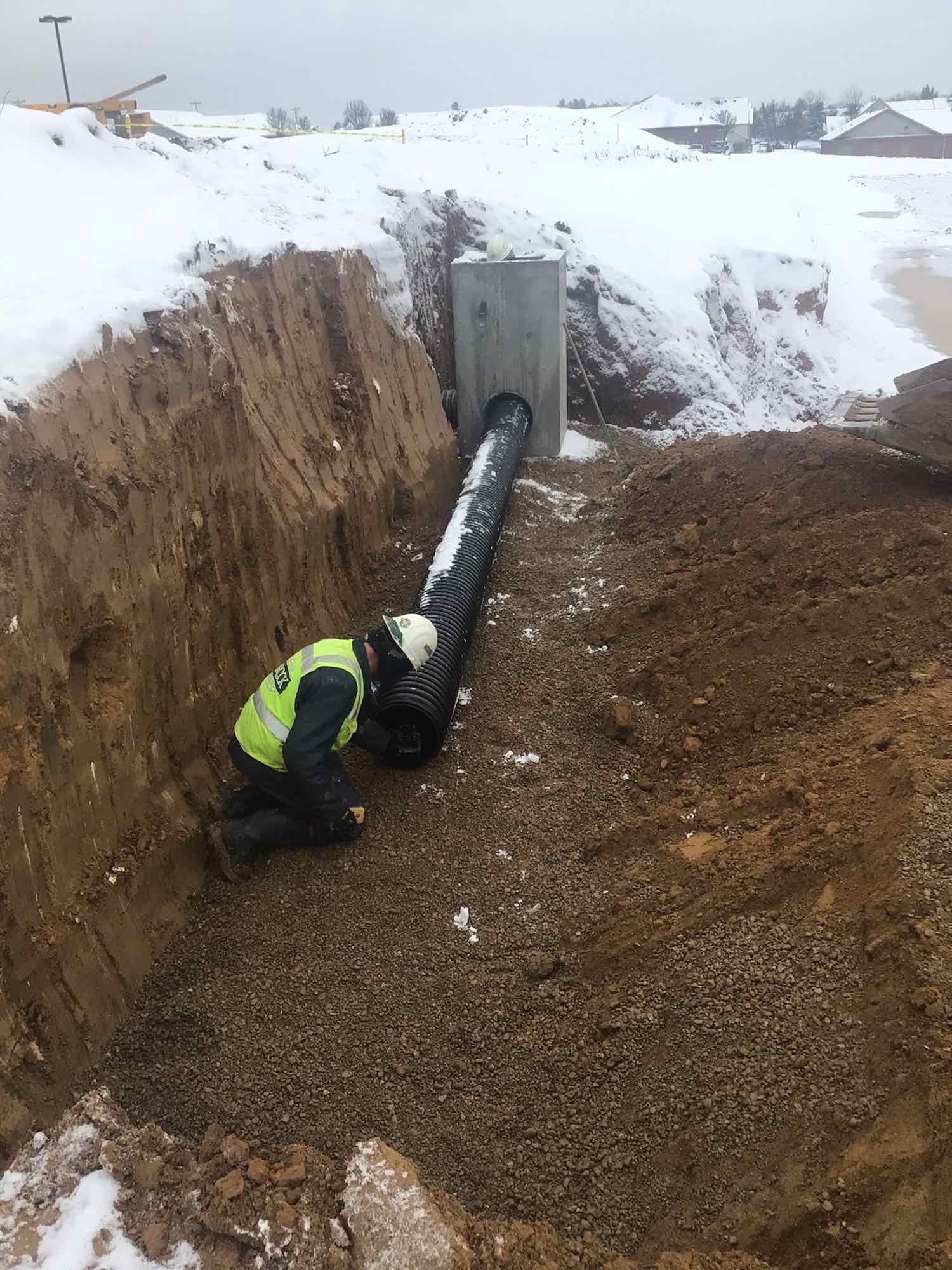 Construction worker in a safety vest working on a drainage pipe in a trench; snowy ground.