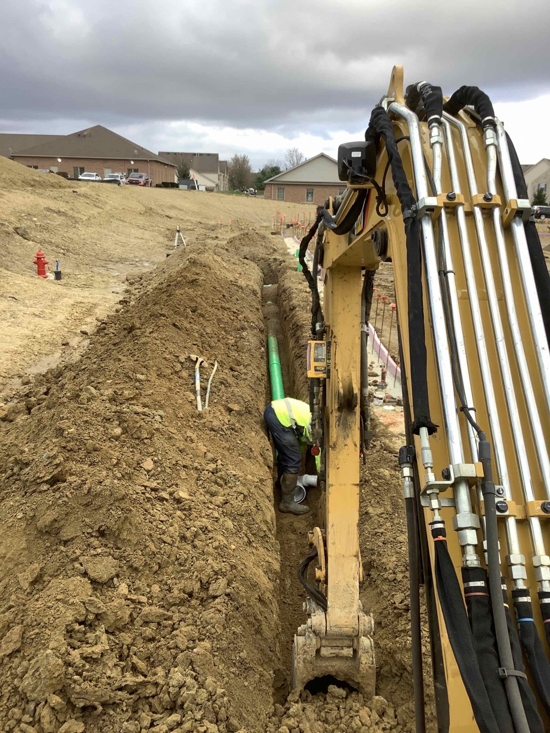 Excavator working near a trench with a worker installing green pipe in a residential area.