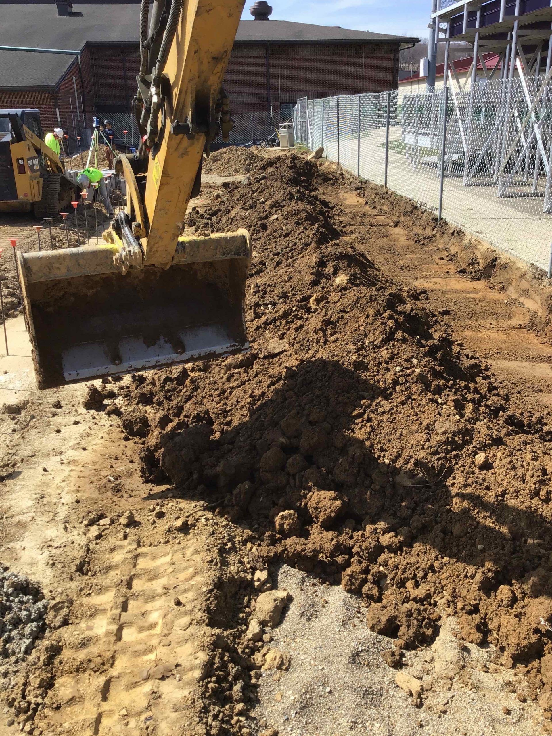 Yellow excavator bucket moving dirt at a construction site. Metal fence around site and brick building in the background. 